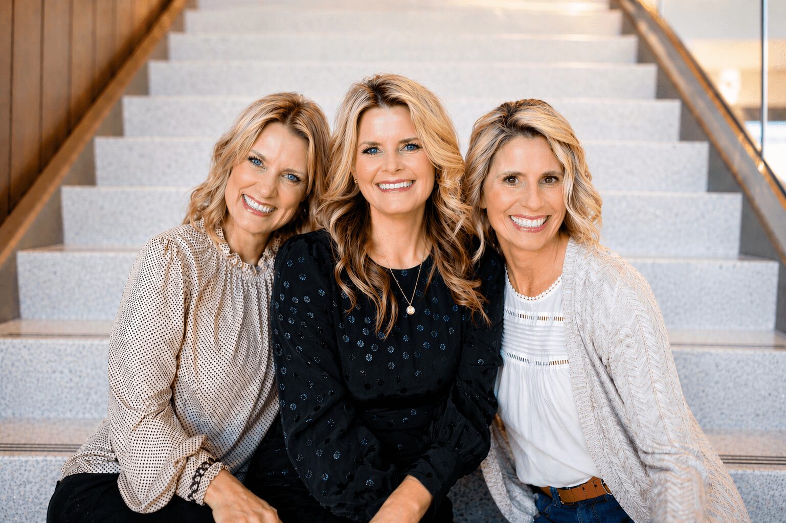 Three women sitting on stairs, smiling, indoors with wooden paneling and railing, casual clothing, blonde hair.