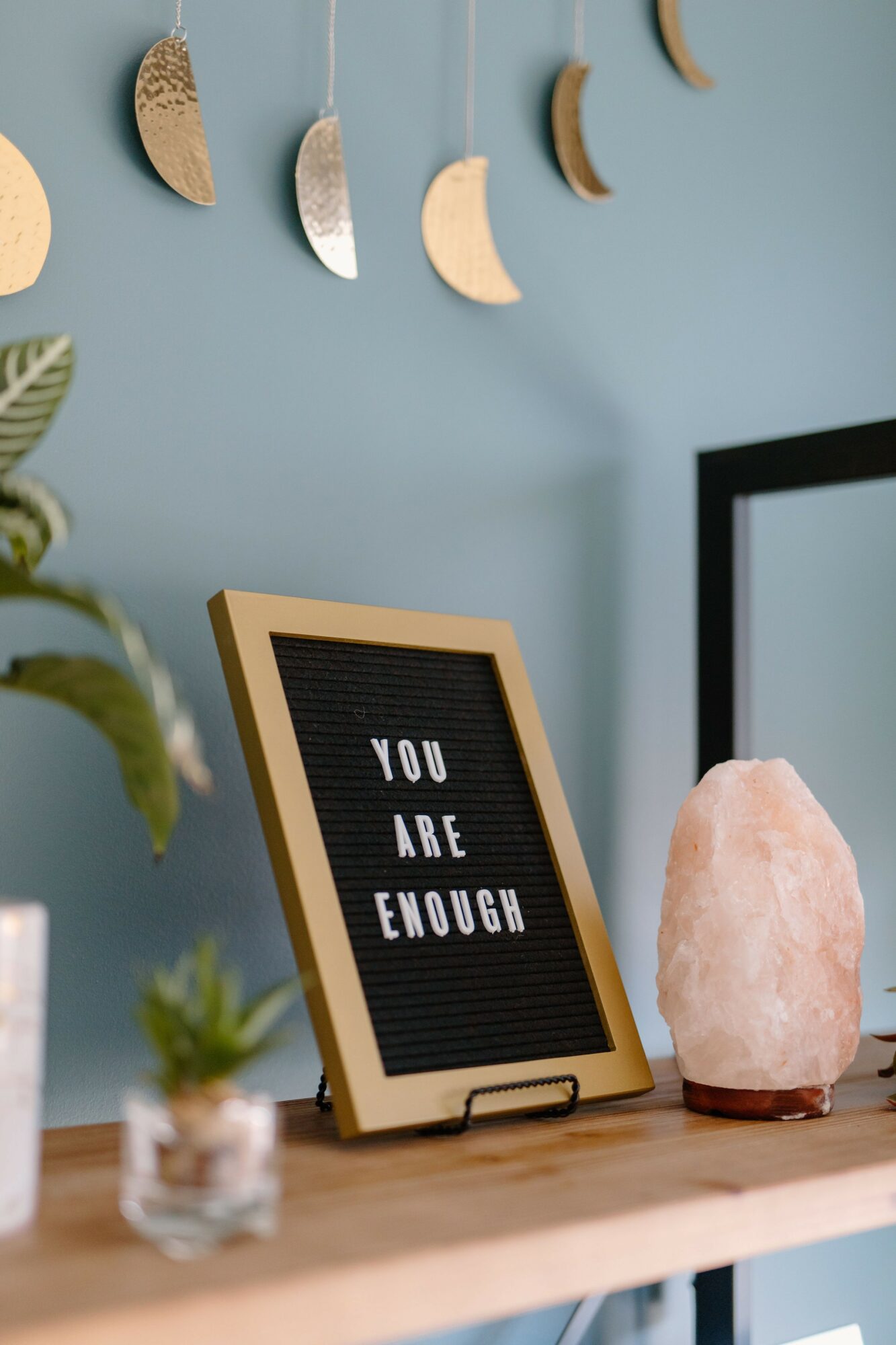 Decorative wall hanging with moon shapes, framed message board with 'You Are Enough', salt lamp, and plants on a wooden surface.