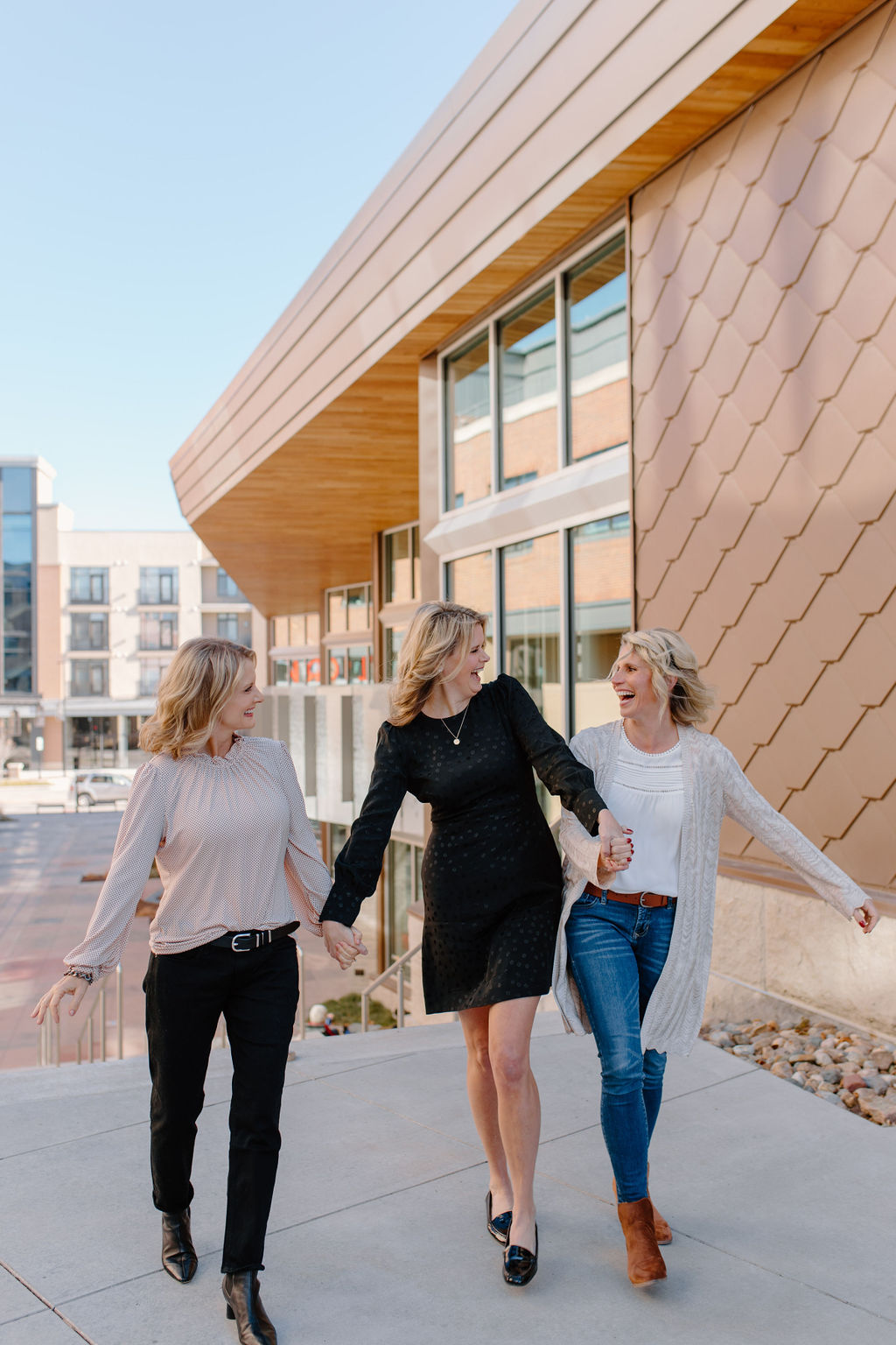 Three women walk together outside a modern building, holding hands and smiling, with a clear sky above.