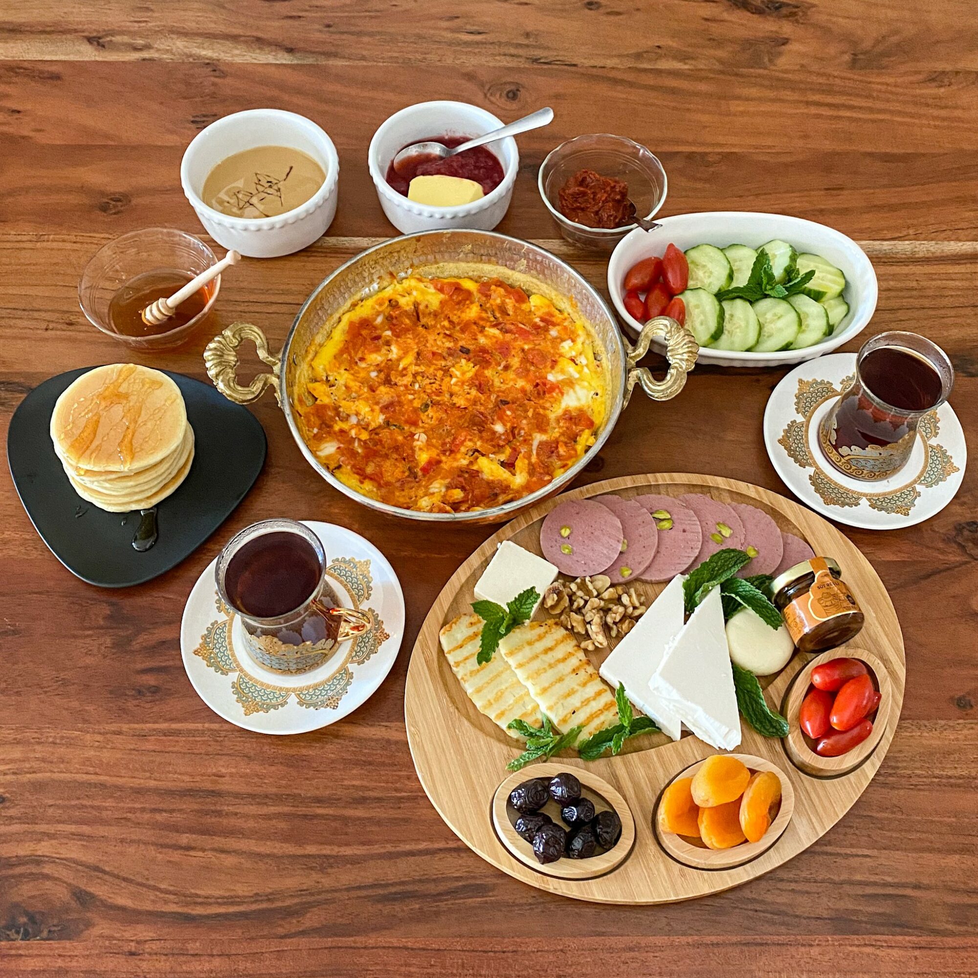 Assorted breakfast foods including a large bowl of scrambled eggs, bread rolls, yogurt with fruit, cucumber slices, tea, and cheese on a wooden table.