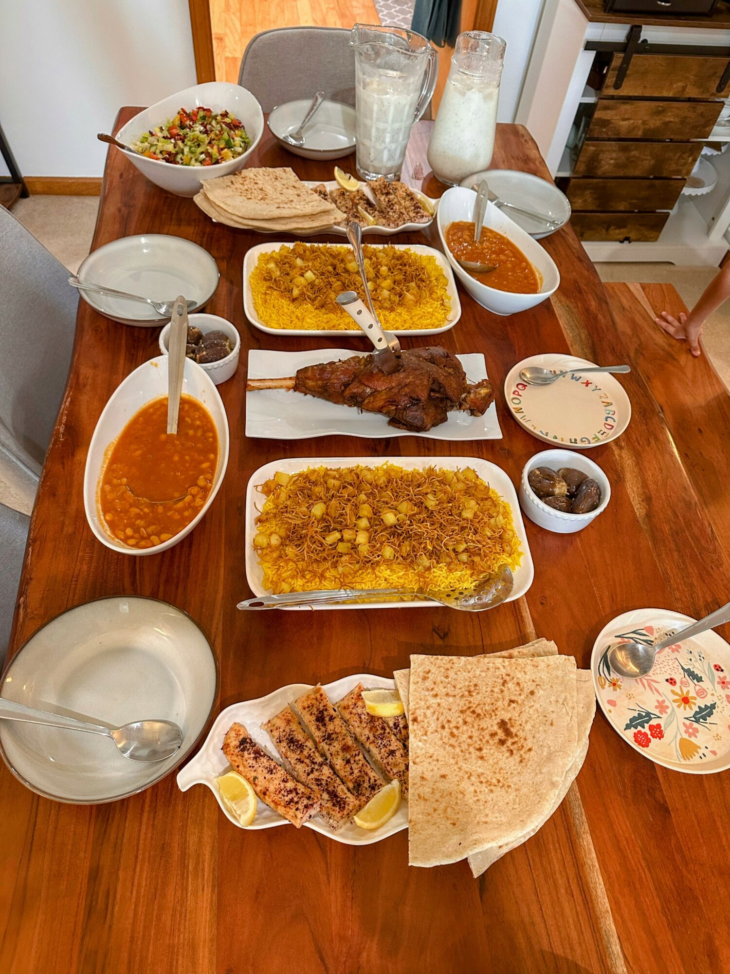 Table with various dishes including rice, bread, grilled meat, beans, salad, drinks, and condiments, set on a wooden table.