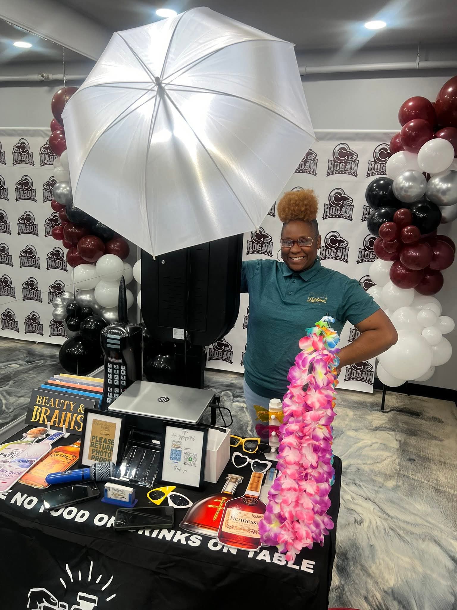 Woman standing behind a table with a large umbrella, balloons, and various products, smiling at the camera.