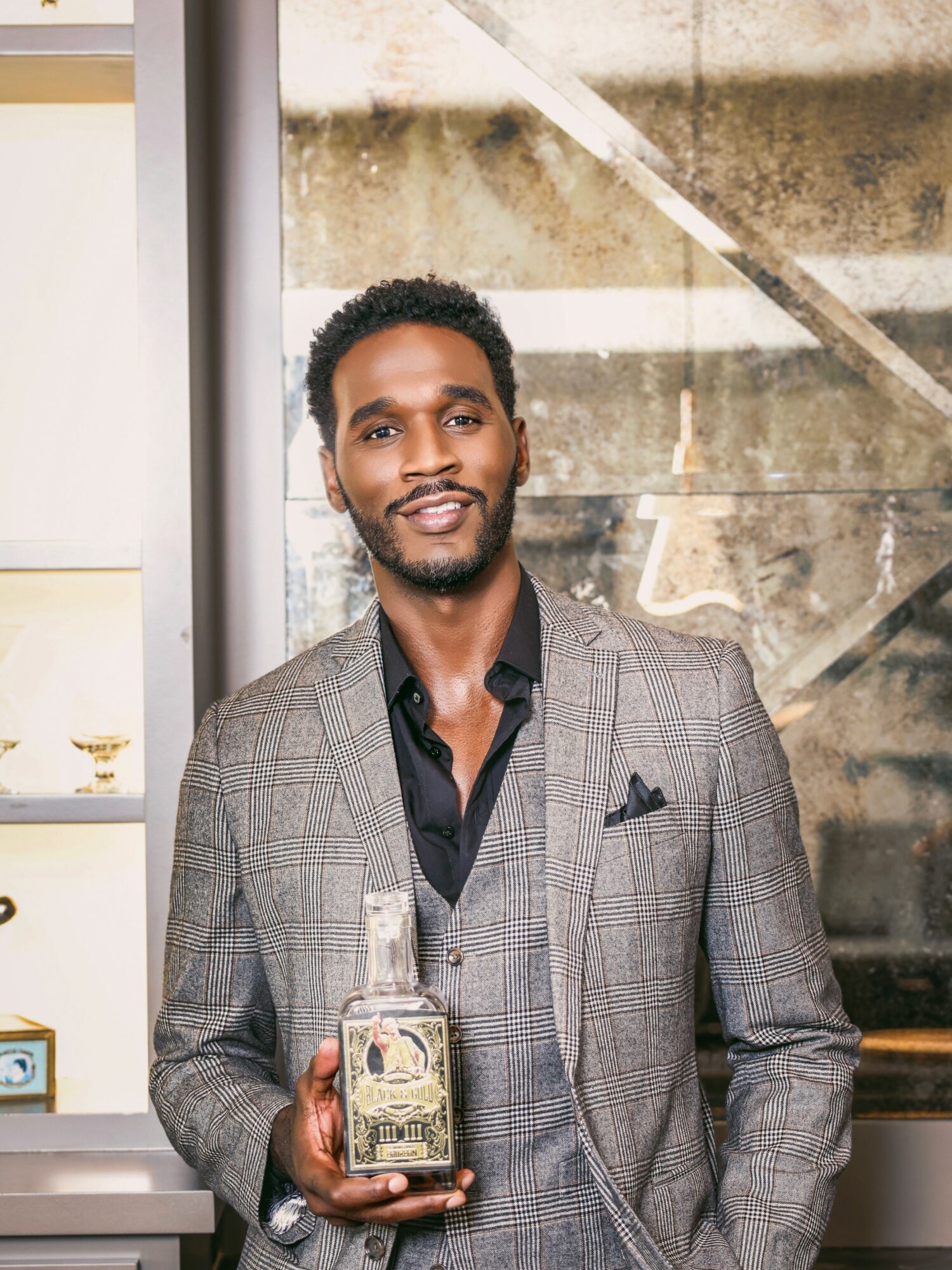 Man in a gray checkered suit holding a glass bottle, standing indoors with stairs in the background.