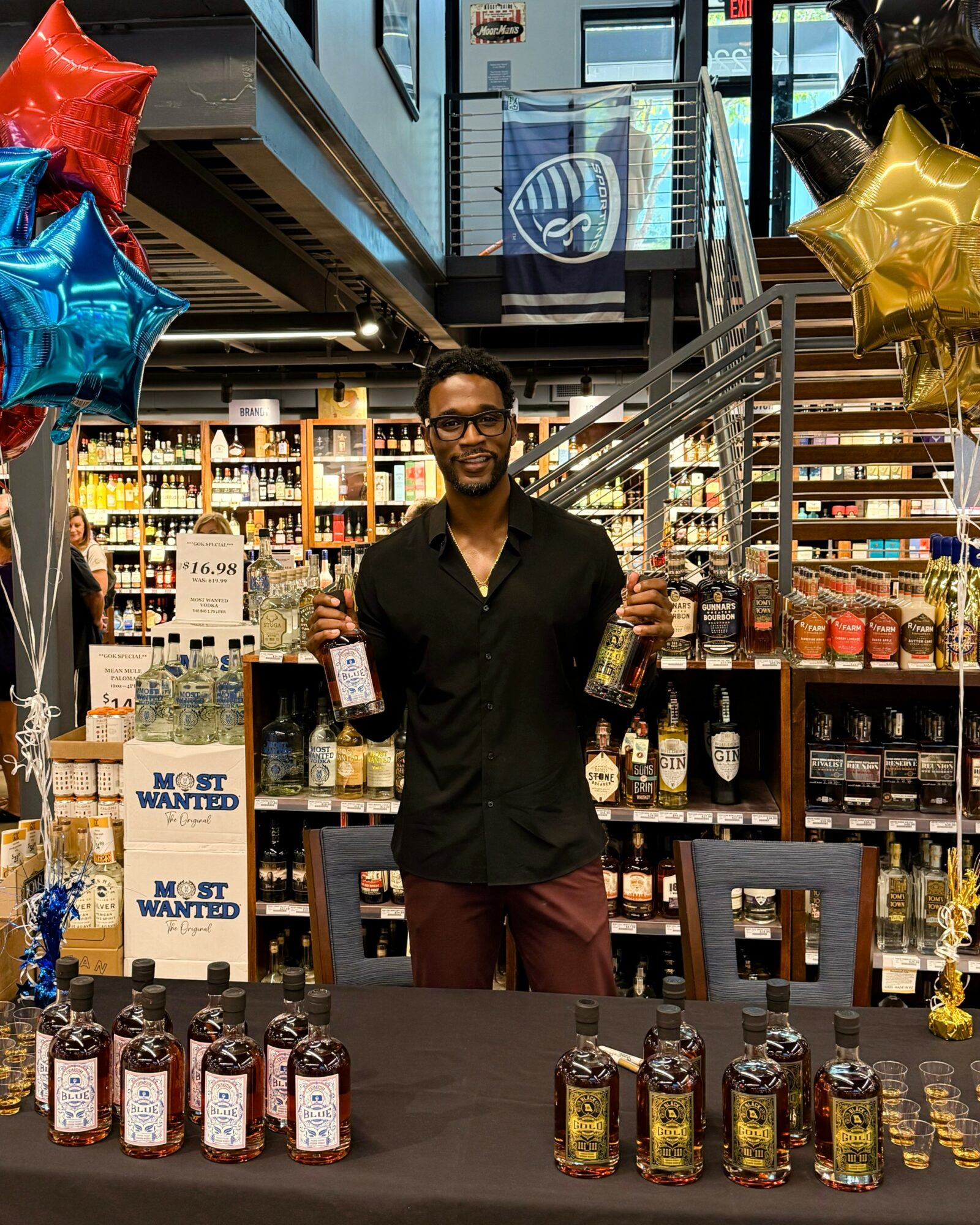 Man standing behind a table with bottles, smiling, in a store decorated with balloons and awards, shelves in background.