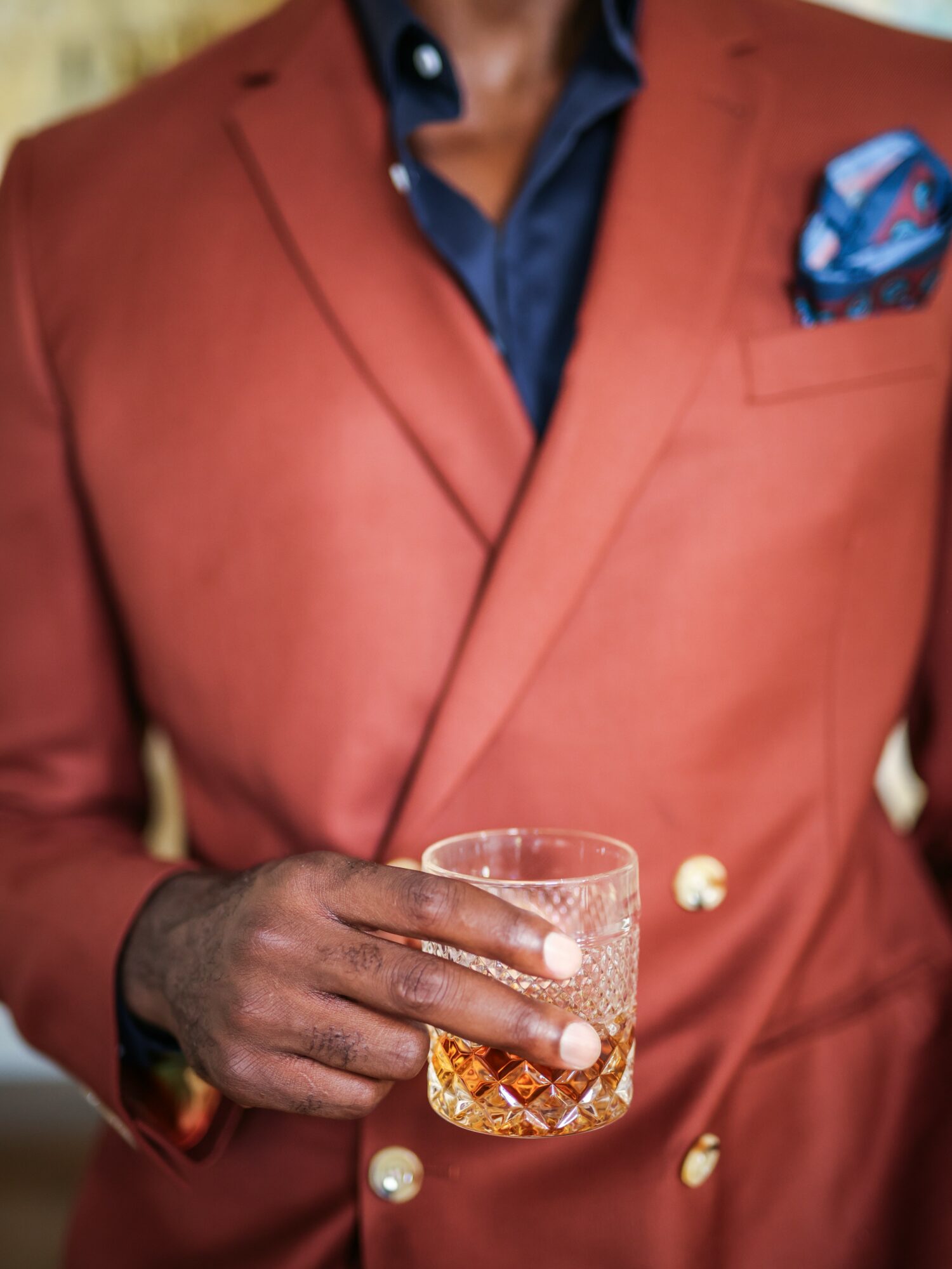 Person in a red blazer holding a glass of amber-colored beverage, with a dark shirt and patterned pocket square.