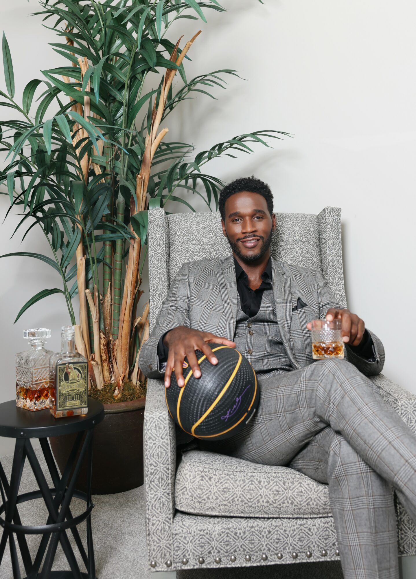 Man in gray suit sitting on a chair, holding a glass, with a basketball and a large plant nearby.