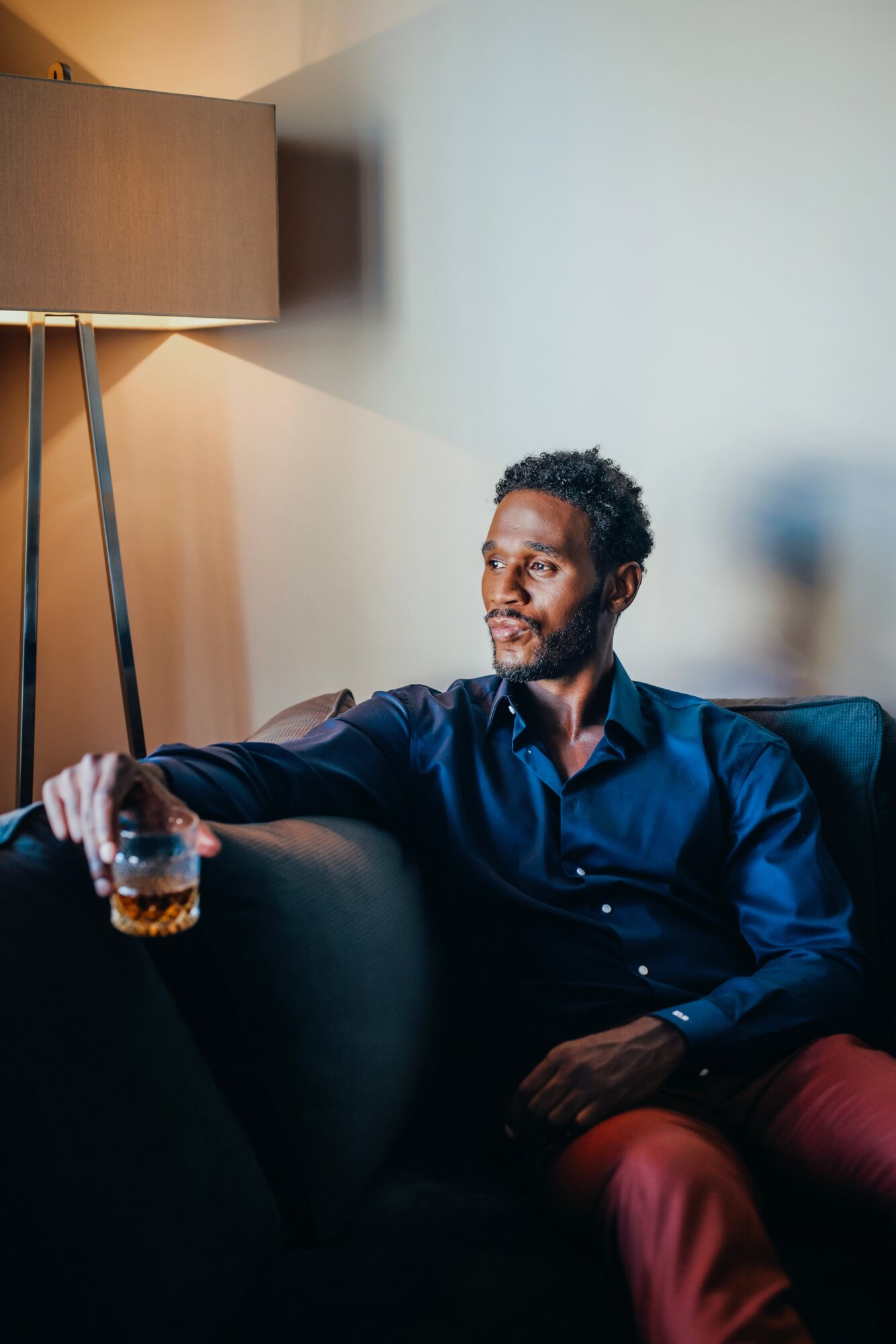 Man with curly hair and beard sitting on a sofa, holding a glass, looking to the side, in a room with a lamp and plain wall.