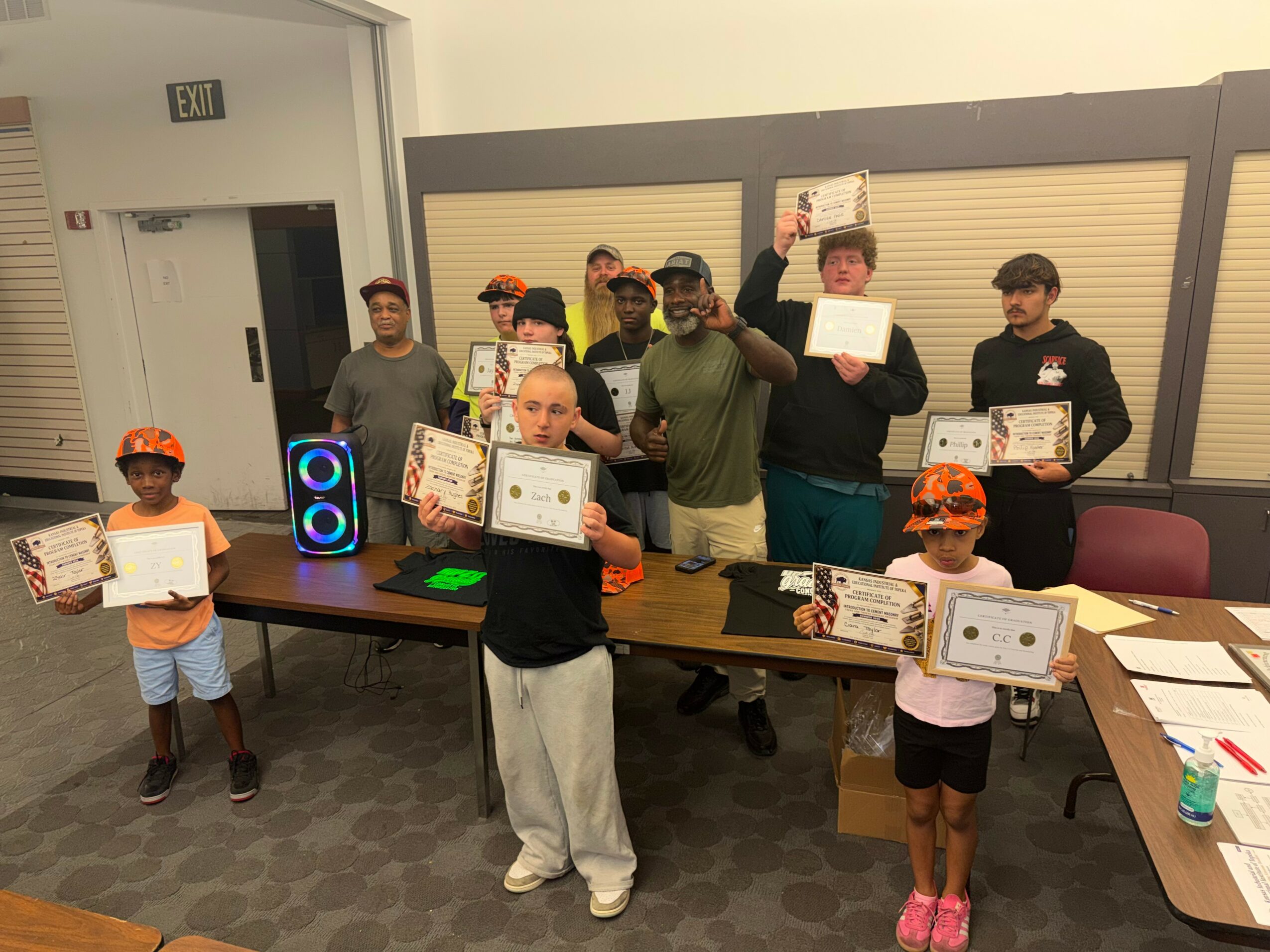 Group of children and adults holding certificates and awards in a room with tables and a speaker, celebrating achievement.
