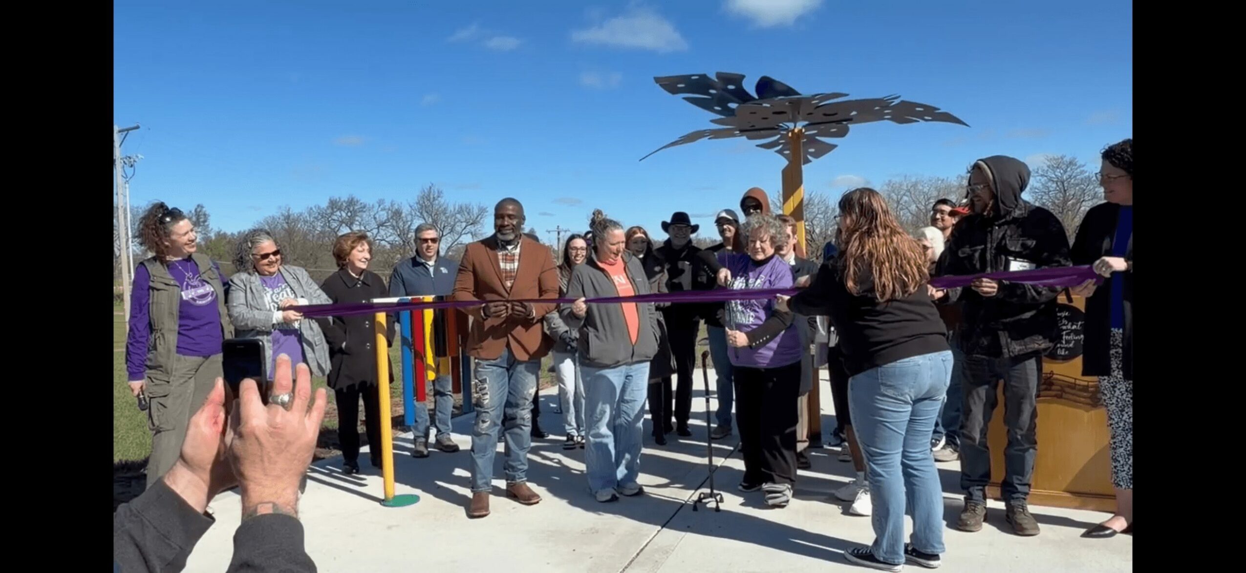 Group of people gathered outdoors near a colorful sculpture, some holding a ribbon, with a clear blue sky.