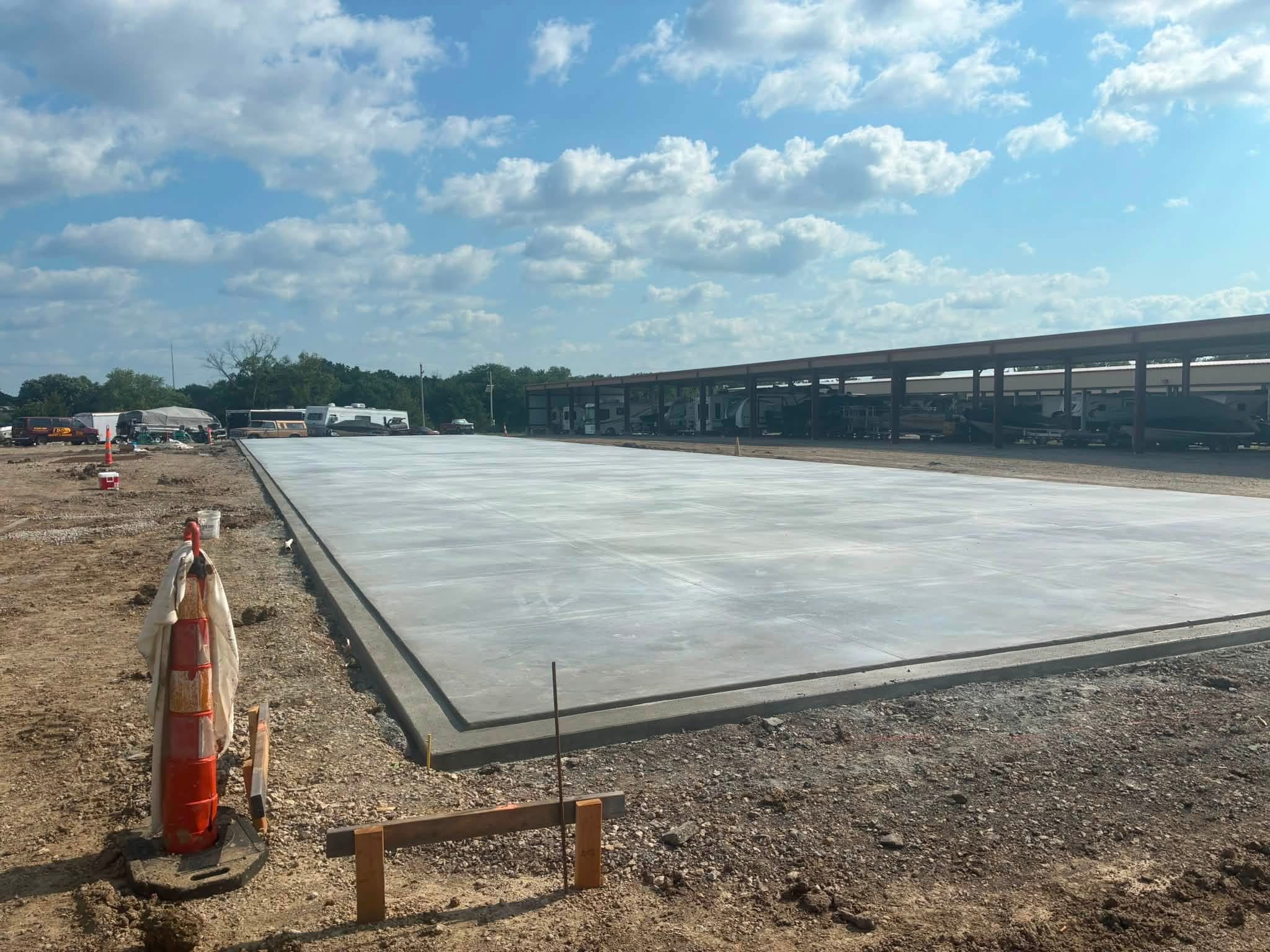 Concrete slab on construction site with orange safety cone and wooden barrier, under a partly cloudy sky.