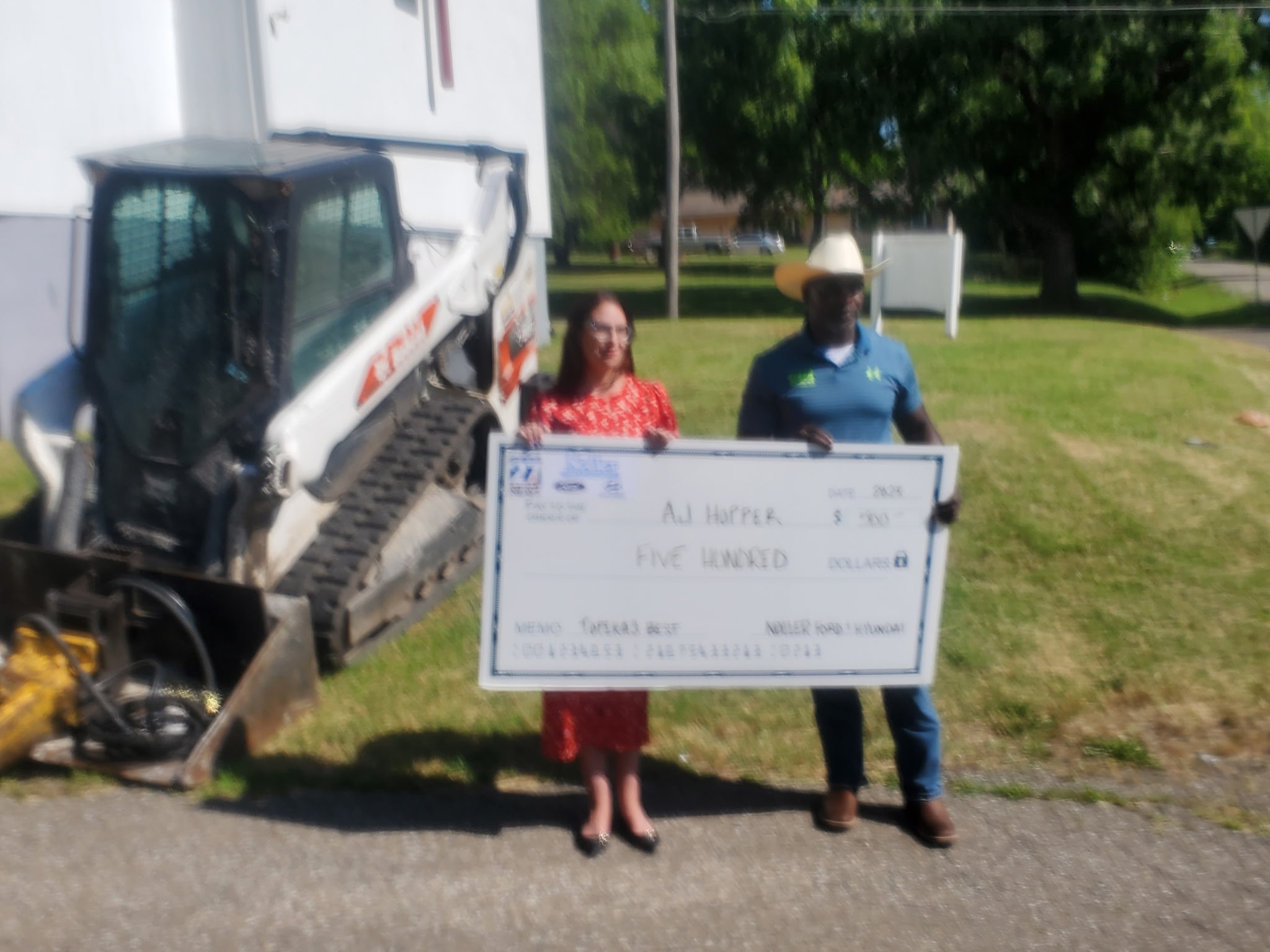 Two people stand outdoors holding a large check, with a construction vehicle nearby and trees in the background.