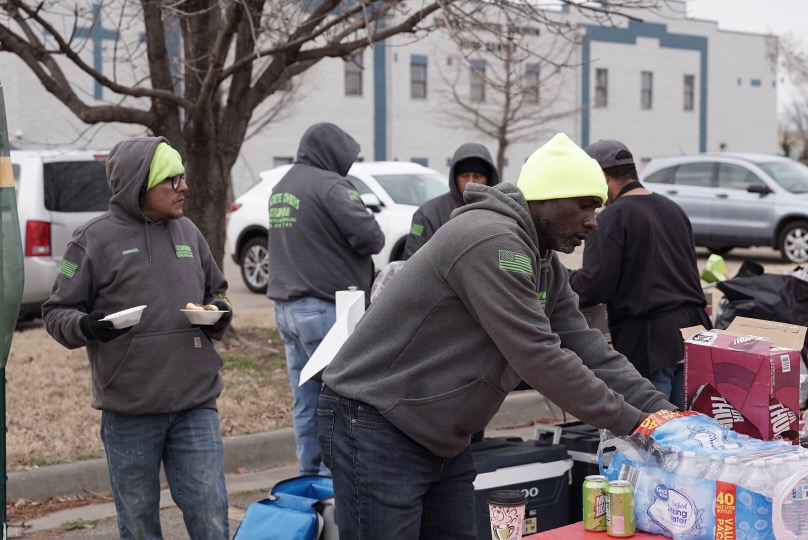 People gather outdoors around tables with supplies, some wearing hoodies and hats, in an urban setting with parked cars.