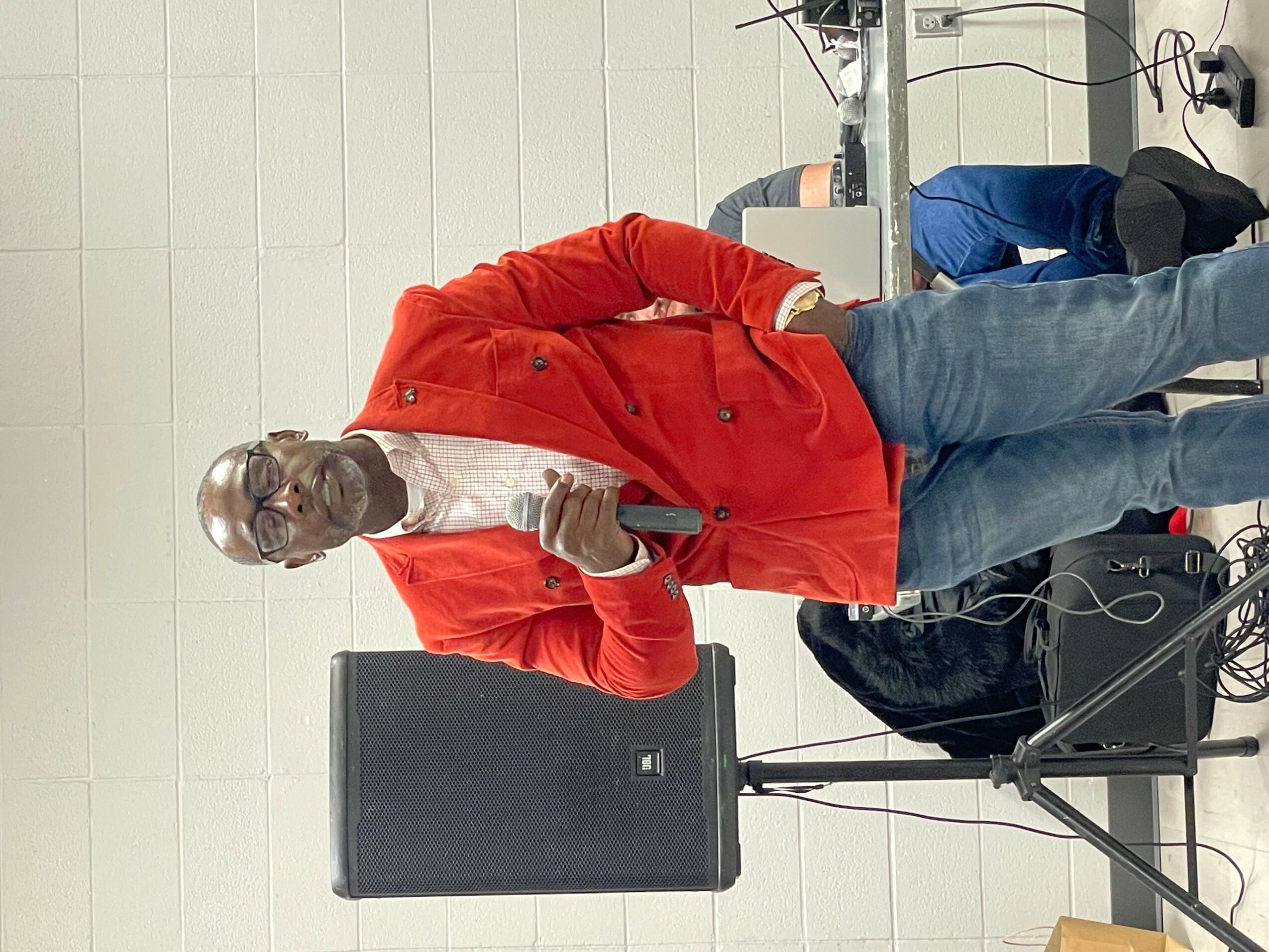 Man in red jacket holding microphone, standing in room with equipment and white tiled wall.