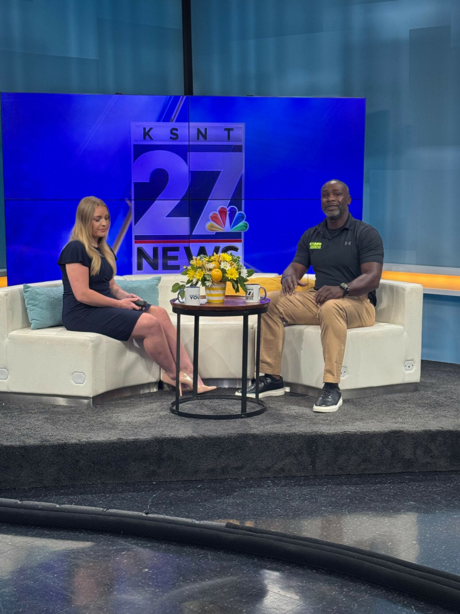Two people sit on a white couch in a studio with a blue background and a large screen displaying 'KSNT 27 NEWS' and a flower arrangement on a small table between them.