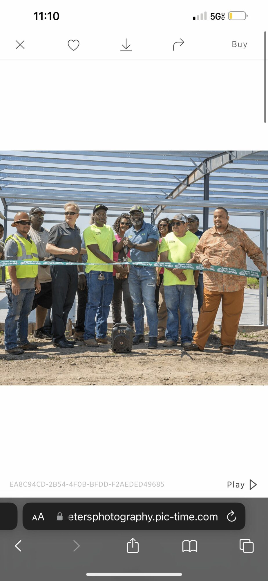 Group of people at a ribbon-cutting event outdoors under a metal roof, some holding scissors, others clapping or smiling.
