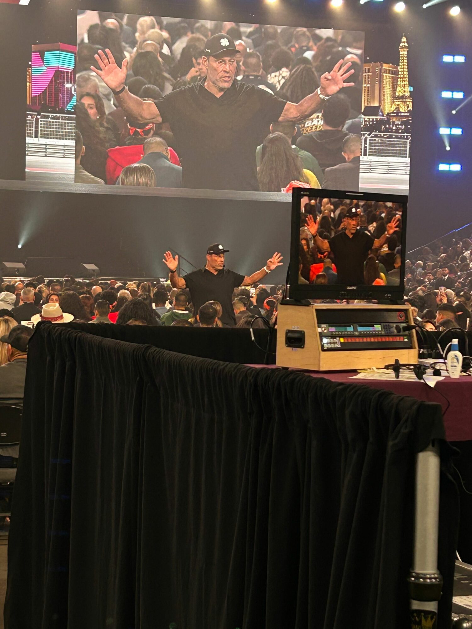 A person on stage with a microphone, audience clapping, and a large screen displaying a crowd scene.
