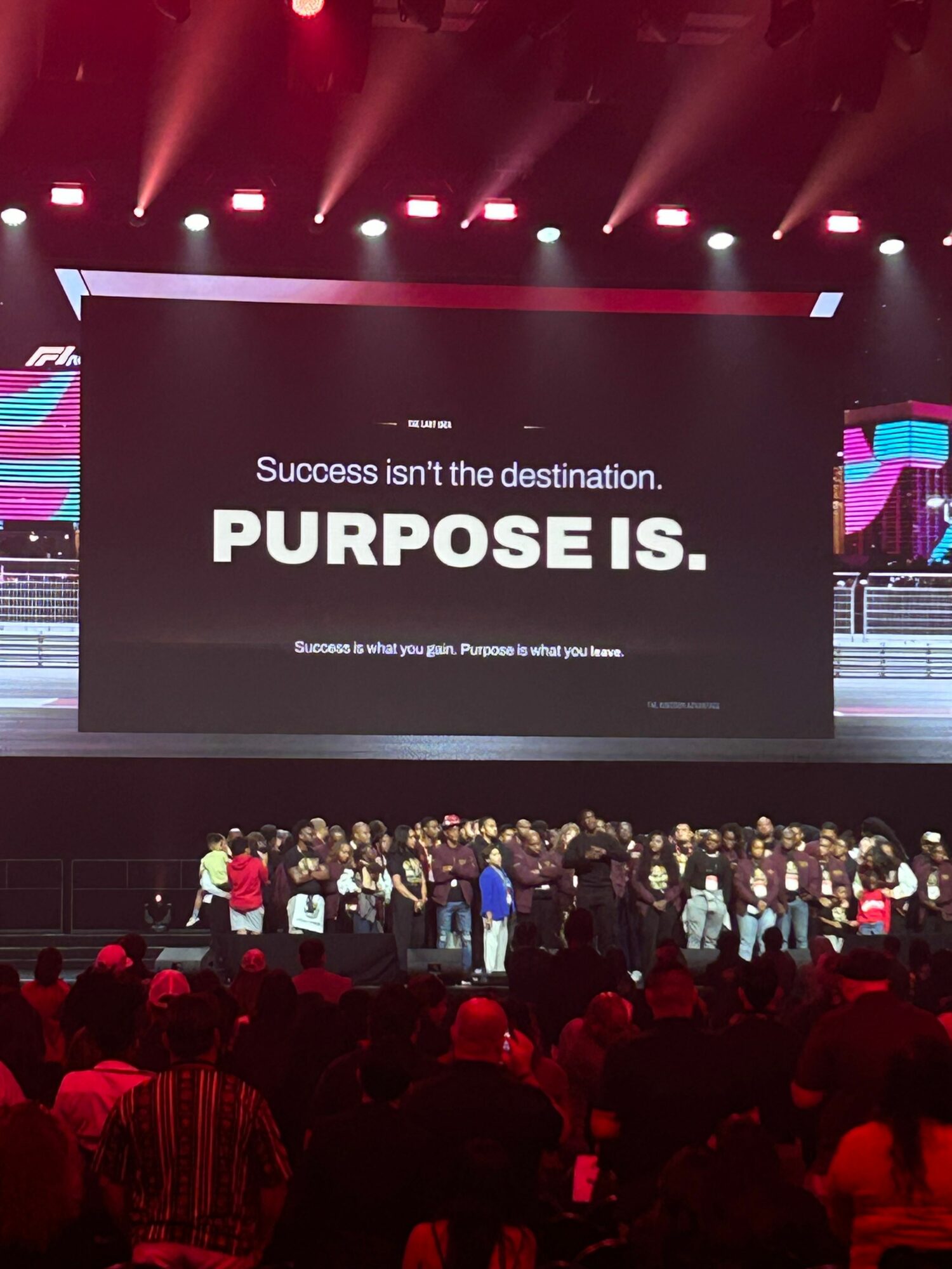 Audience watching a presentation with a large screen displaying the words 'Success invites destination. PURPOSE IS.' in a dark room with red lighting.