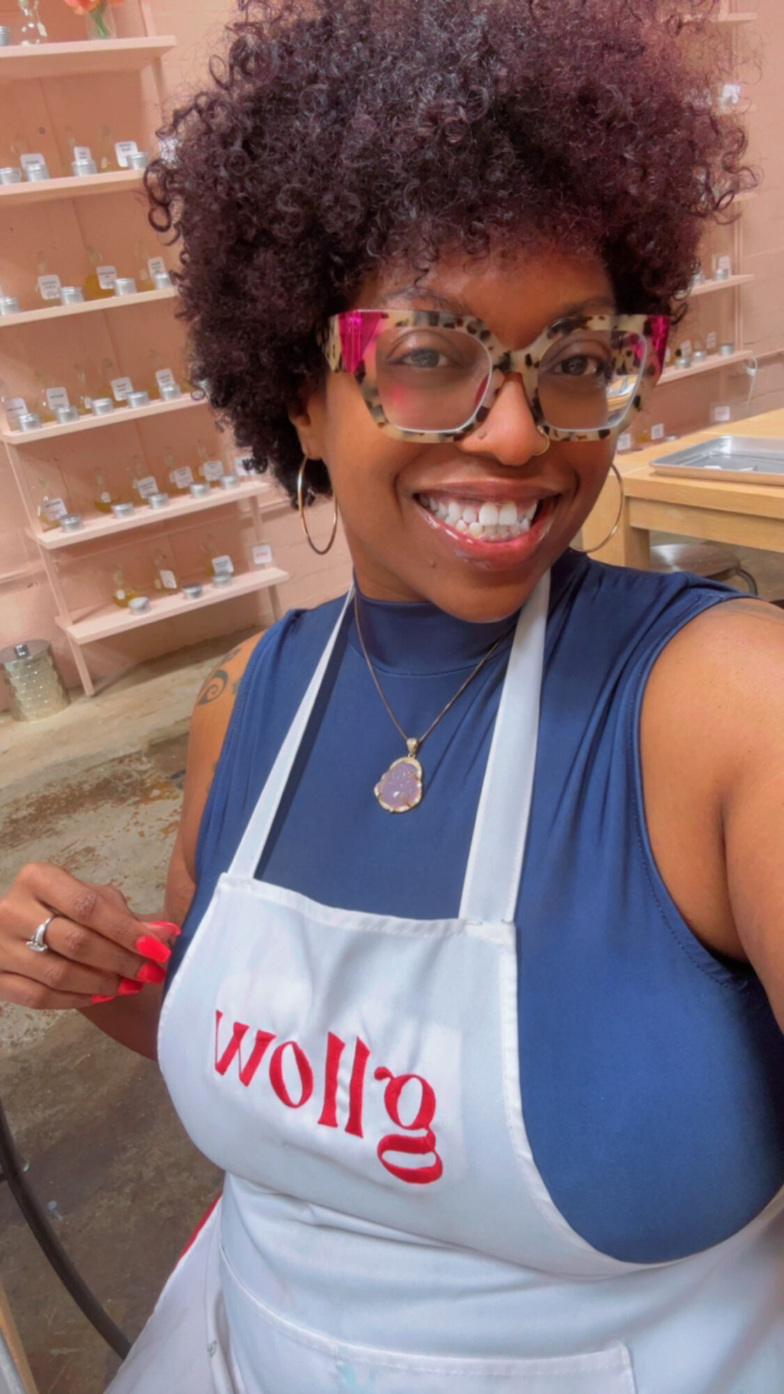 Smiling woman with curly hair wearing glasses and a blue shirt, in a store with shelves in background.
