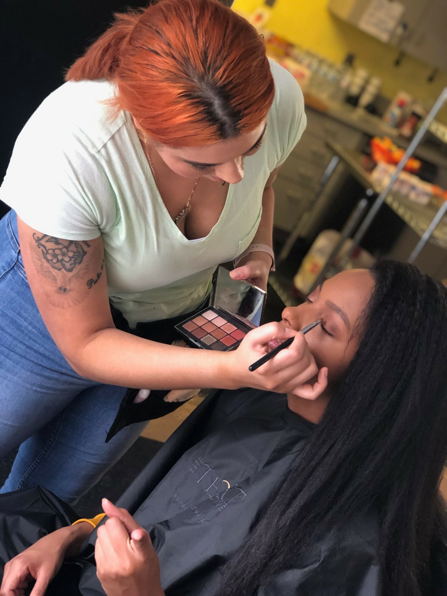 Makeup artist applying makeup to a woman in a salon, holding a palette and brush, with shelves in the background.