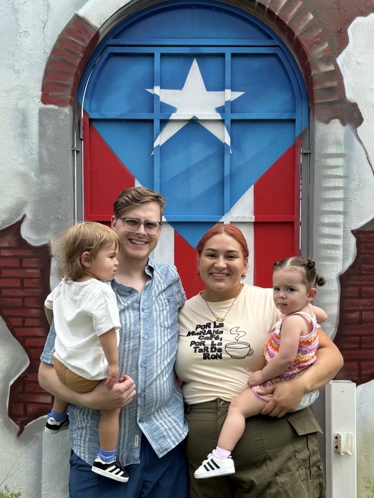 Two women holding children stand in front of a colorful door with a star and flag design, smiling outdoors.