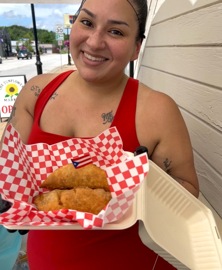 Smiling woman in red top holding fried food in checkered paper, standing outside near a white wall.