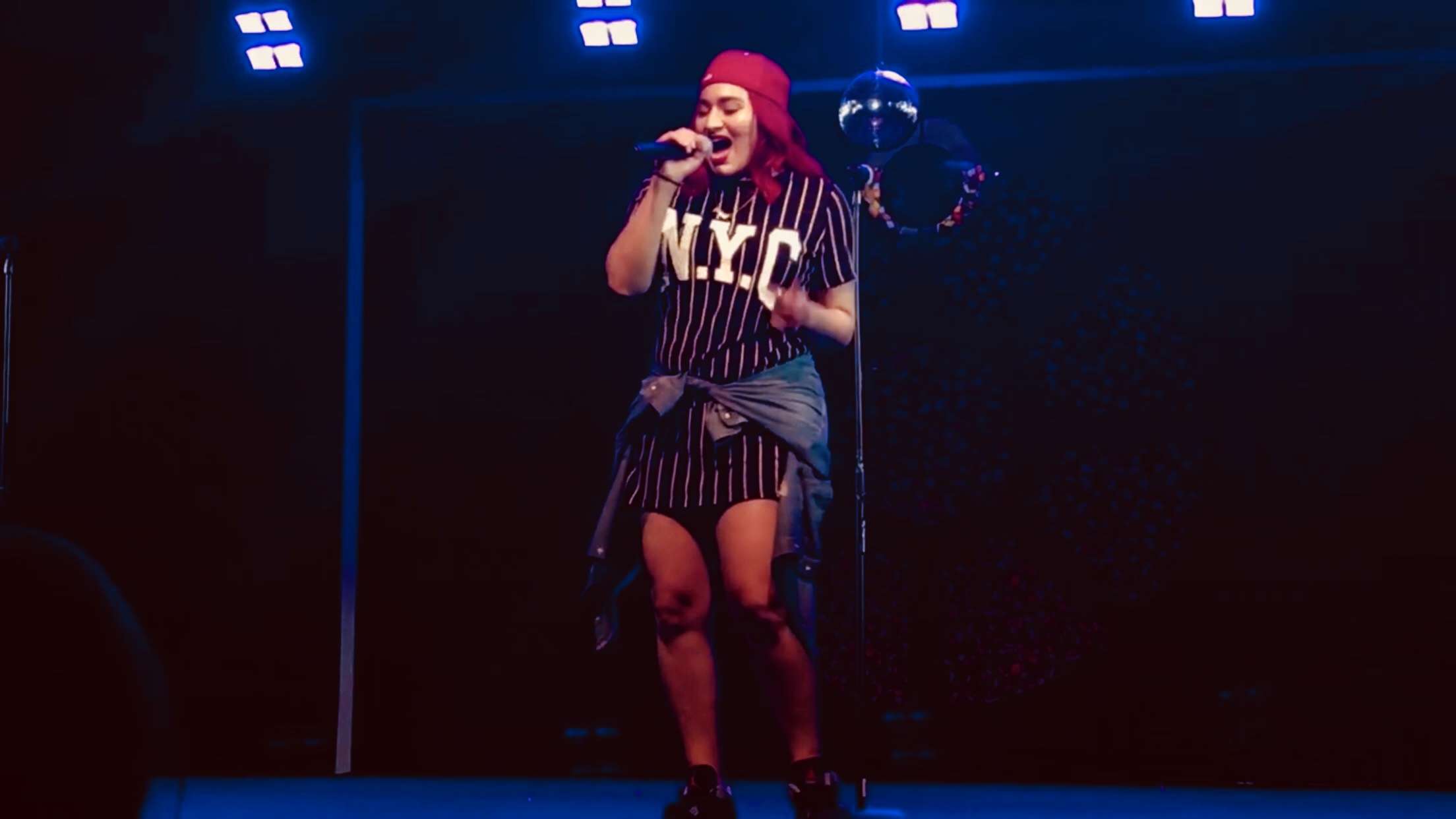 Performer singing on stage with bright lights, wearing a striped outfit and red head covering.