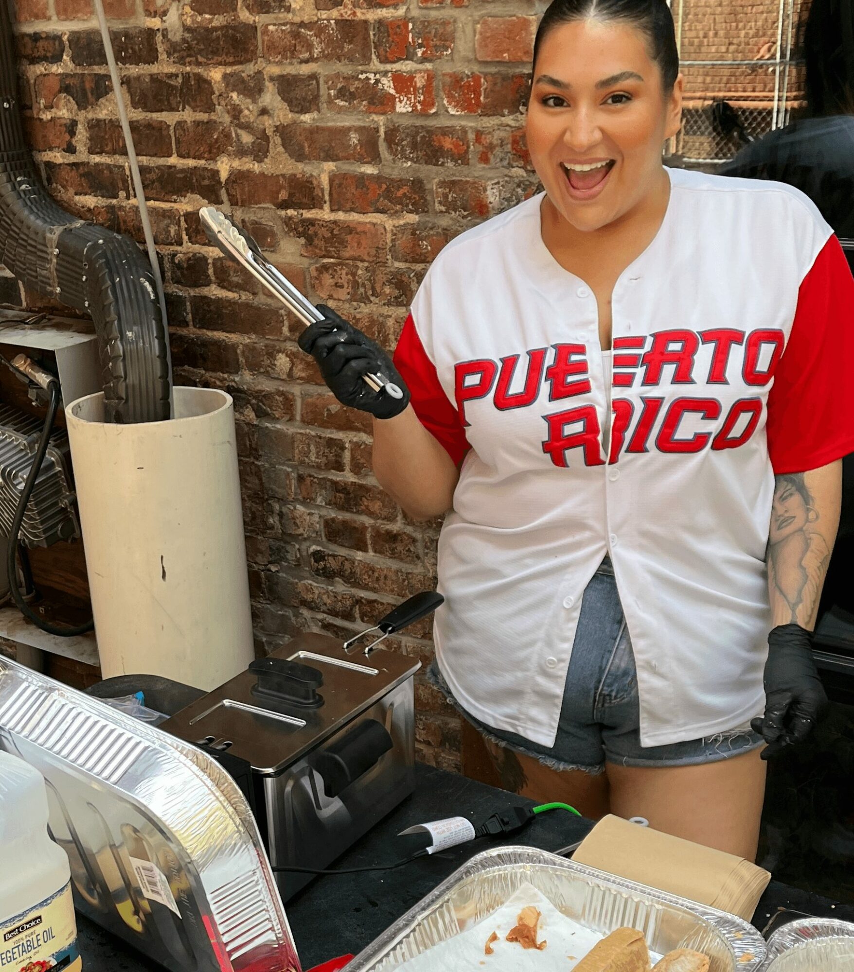 Woman smiling, holding tongs, standing behind a food counter with a brick wall background.