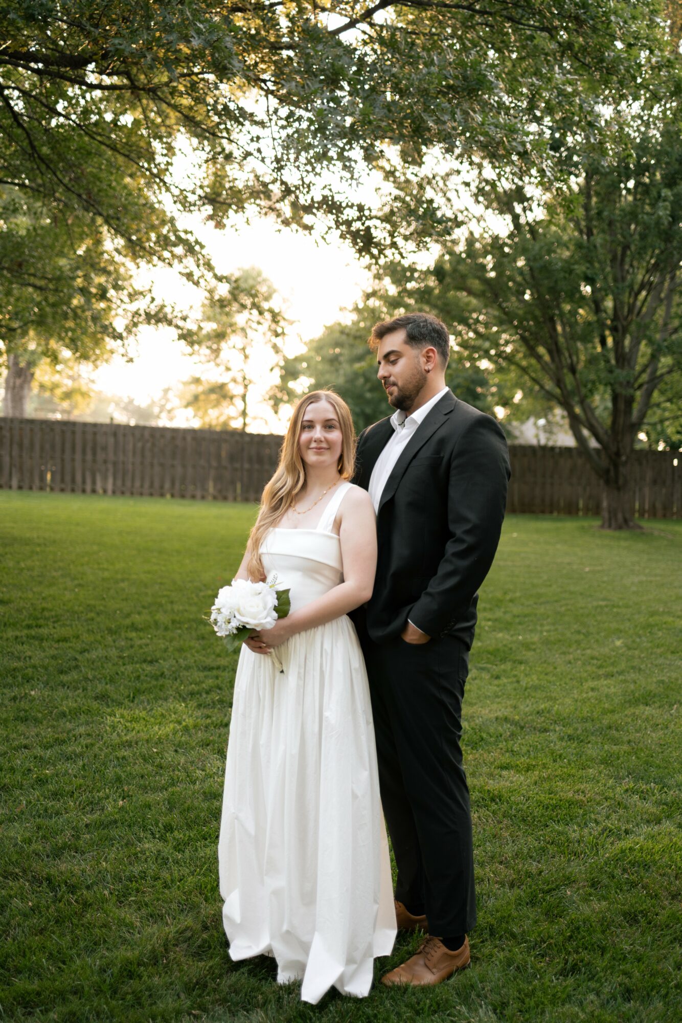 A bride and groom standing outdoors on grass with trees in the background, smiling, during daytime.