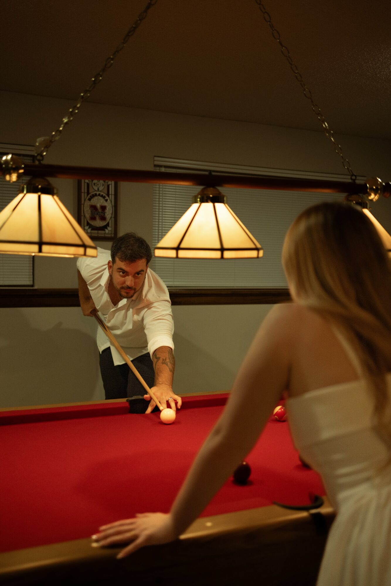 Man aiming cue at pool ball on red pool table, woman watching, hanging lights above, indoor setting.