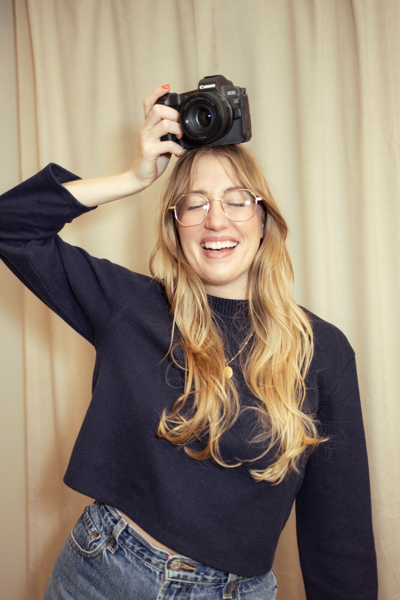 Young woman with glasses smiling, holding a camera above her head, standing in front of a beige curtain.