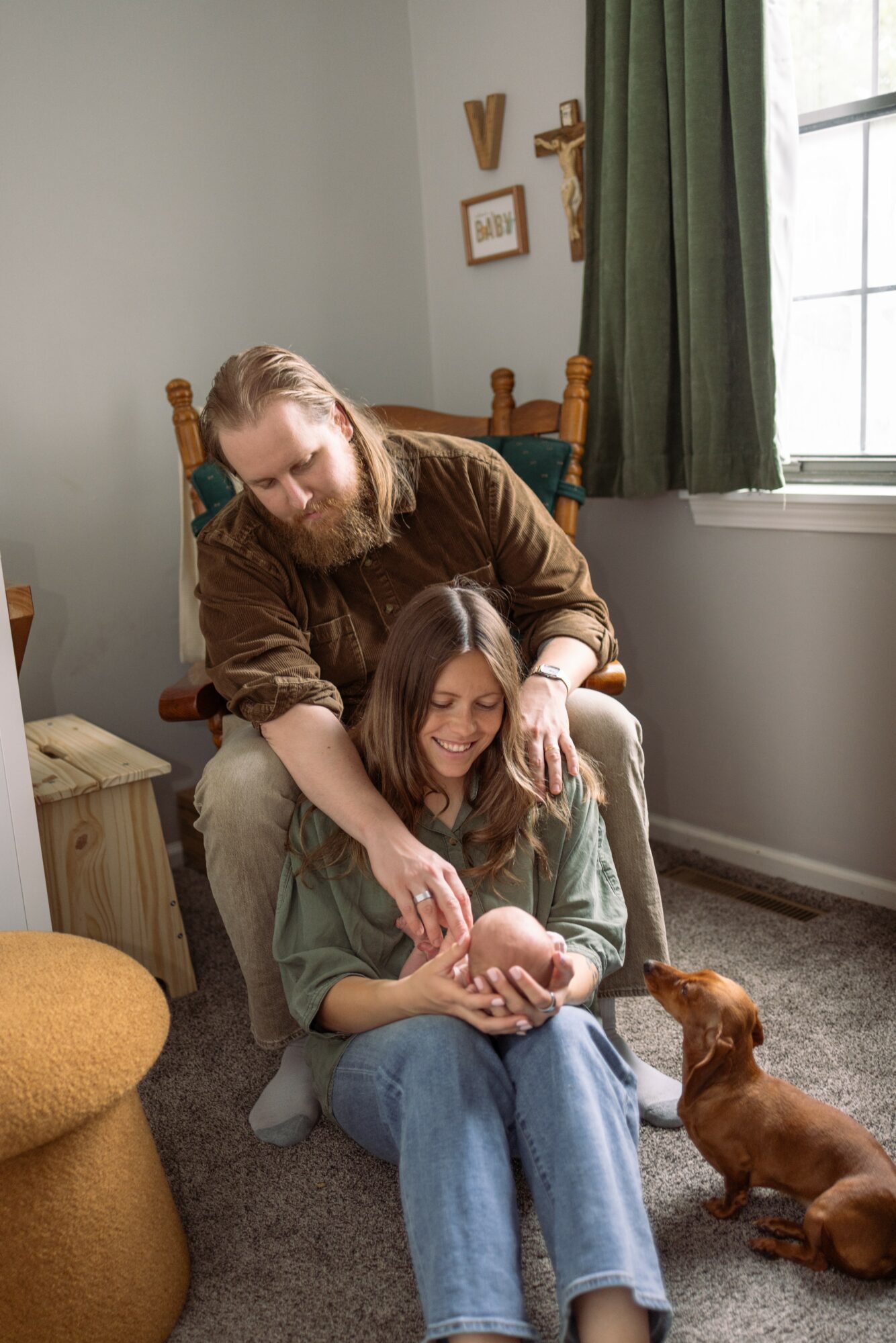 A woman sitting on a chair holding a baby, with a girl and a dog nearby in a cozy room.