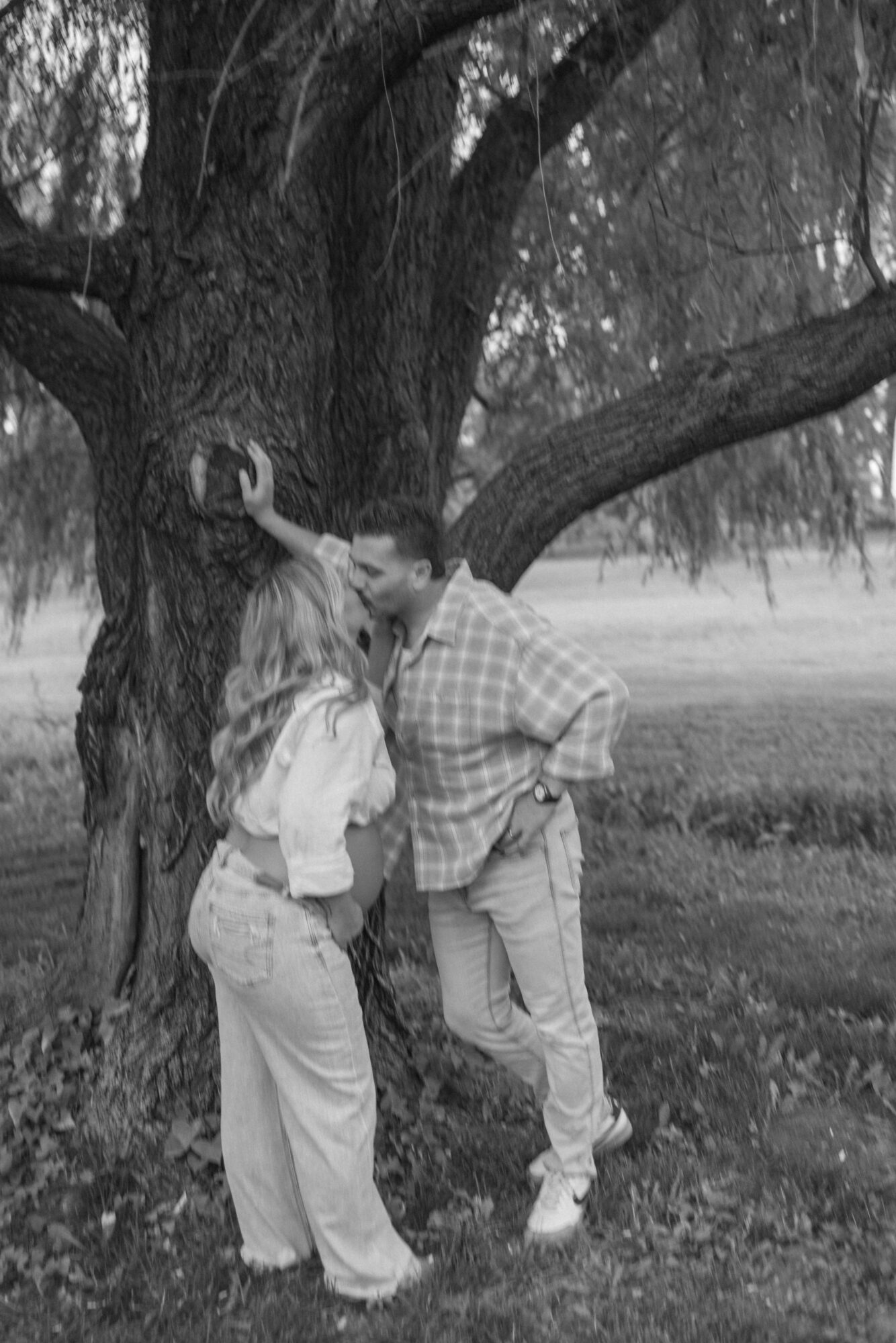 Two people stand near a large tree, facing each other, outdoors in a park-like setting, black and white photo.