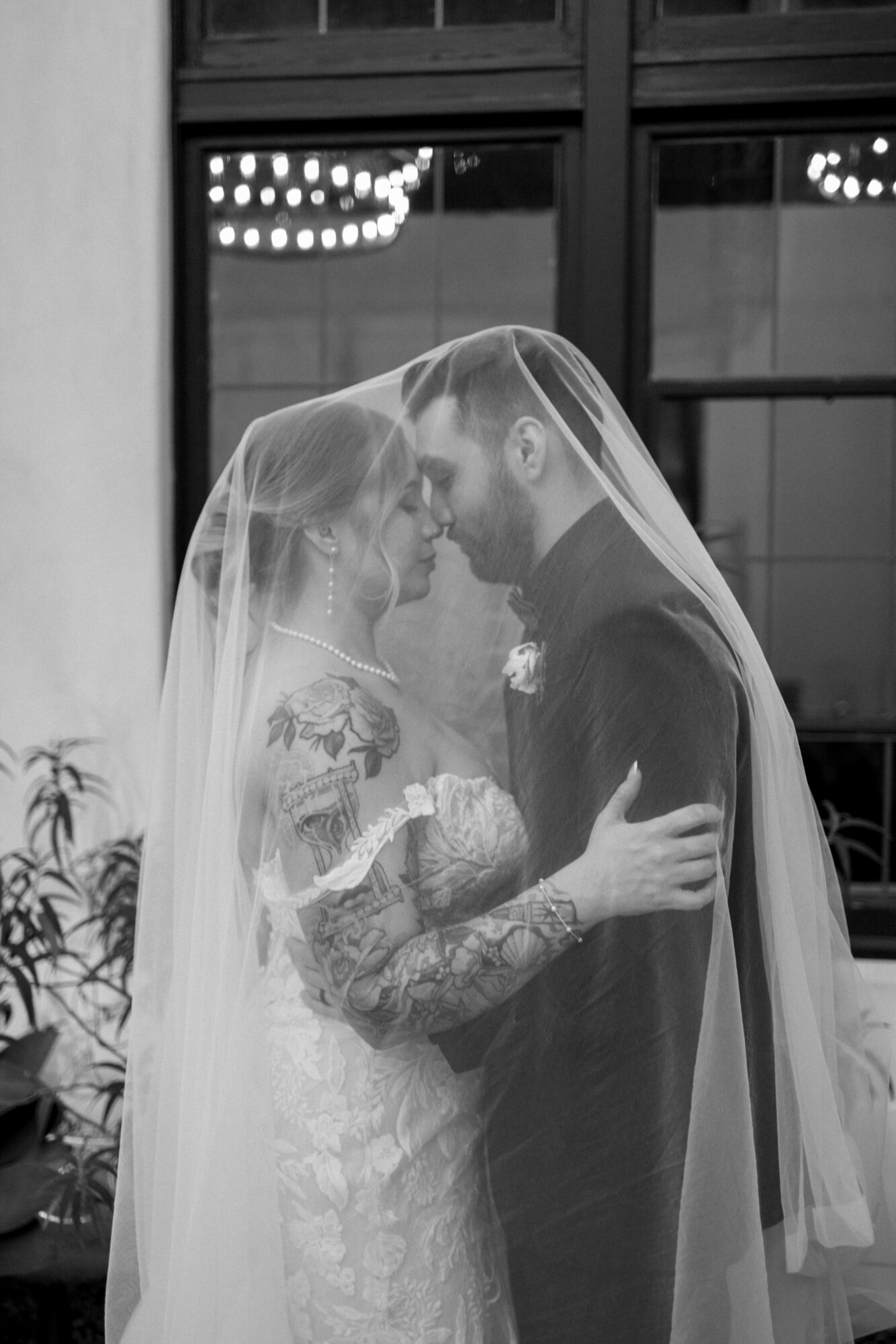 Bride and groom face each other under a veil, holding each other closely, in front of a window with lights.