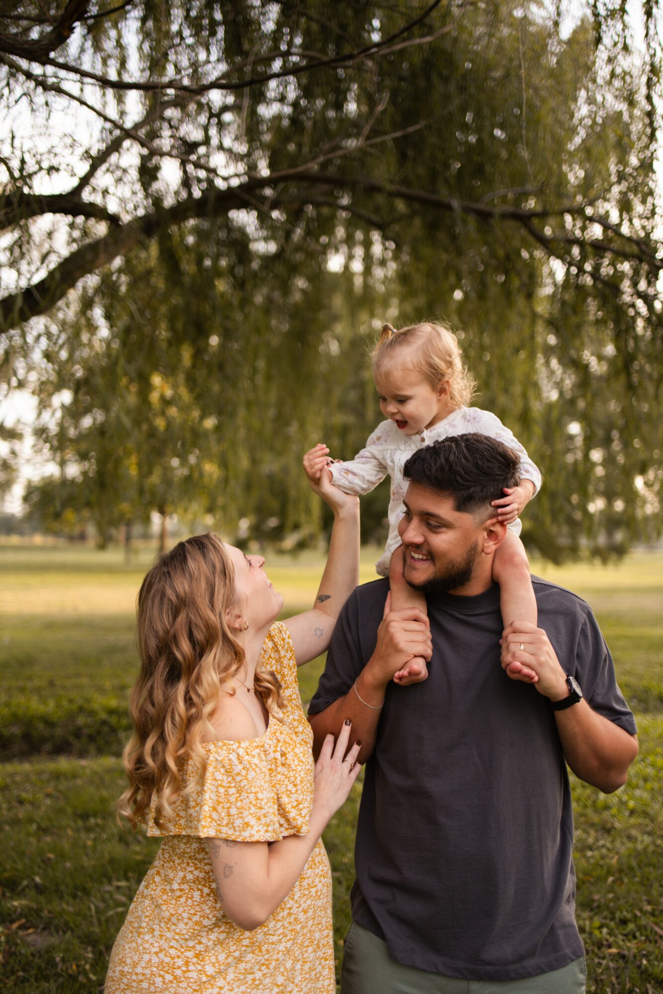 A family of three outdoors under a large tree, with the father carrying a young girl on his shoulders and the mother reaching up to her.