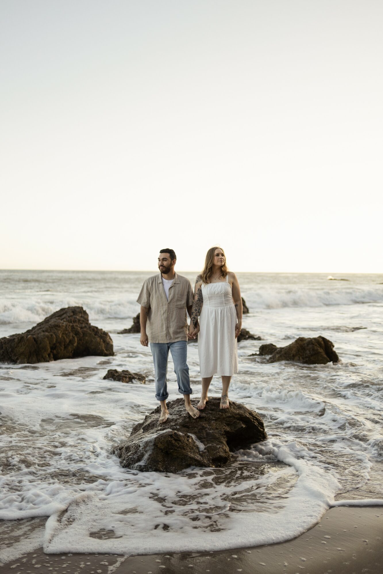 A couple stands on rocks at the beach with ocean waves behind them, under a clear sky.