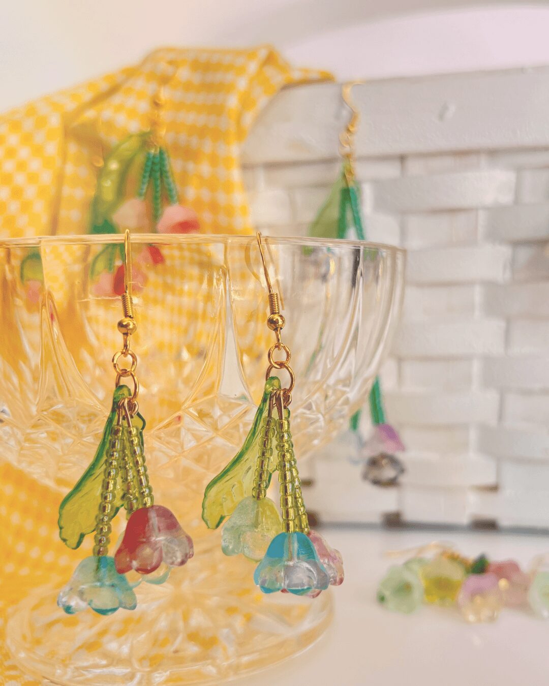 Colorful beaded earrings with flower-shaped charms hanging from a glass bowl, with a yellow checkered cloth and white basket in background.
