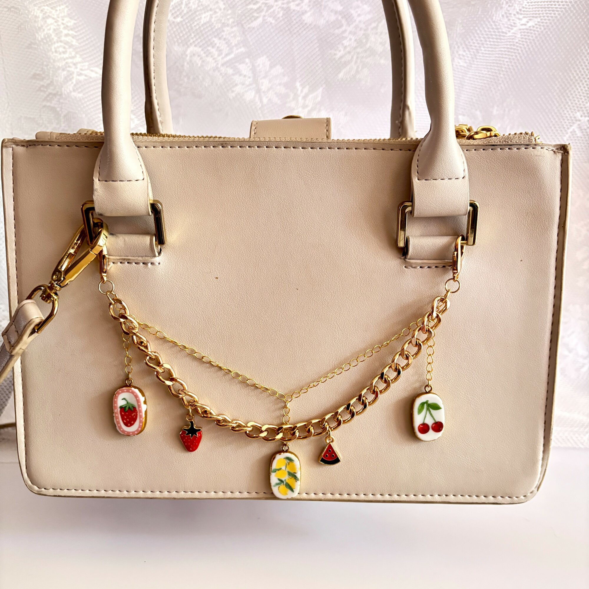 Cream-colored handbag with gold chain and colorful charms hanging from it, against a light background.