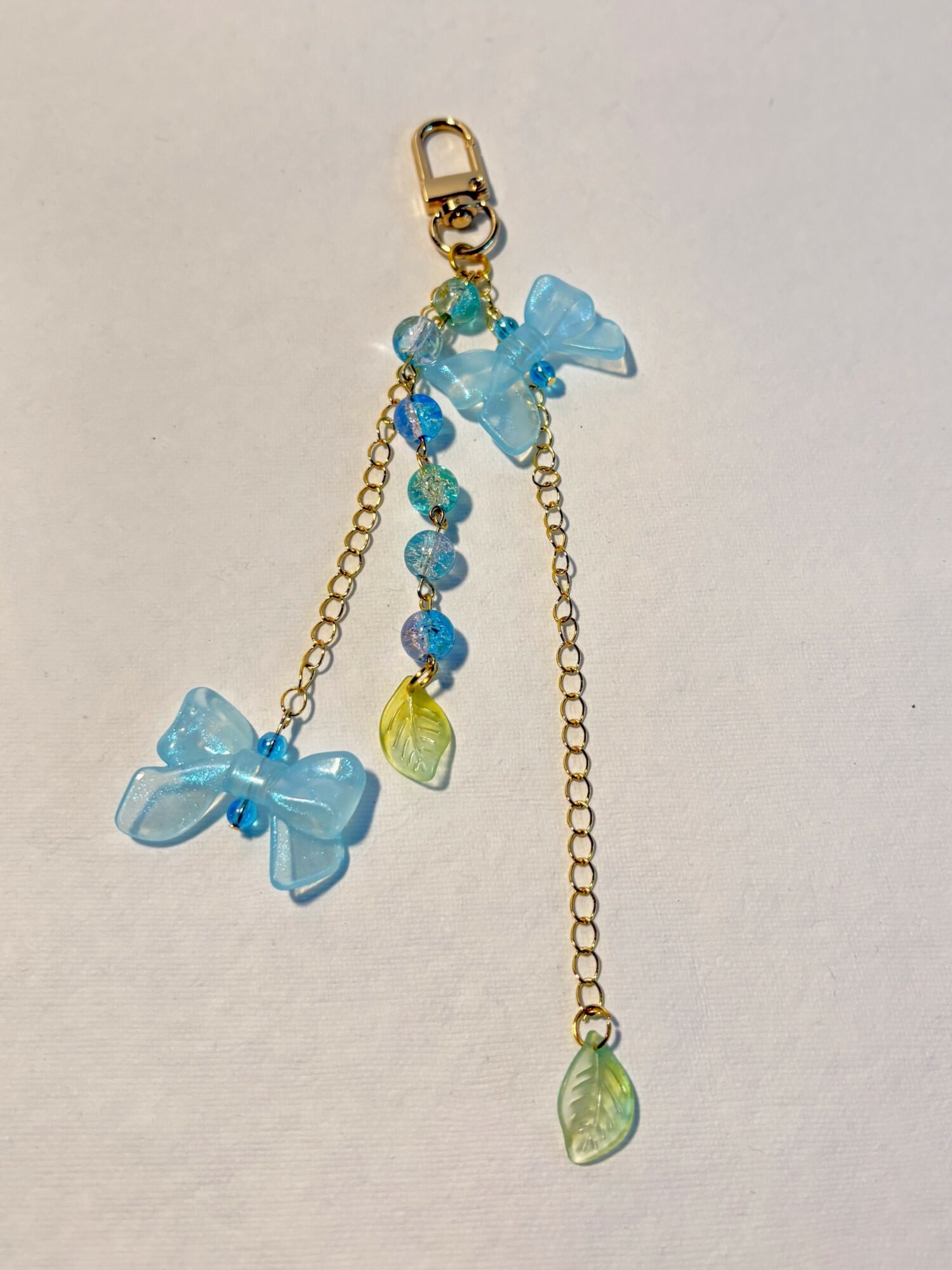 Gold chain with blue bows and beads, and green teardrop charms, hanging against a plain background.