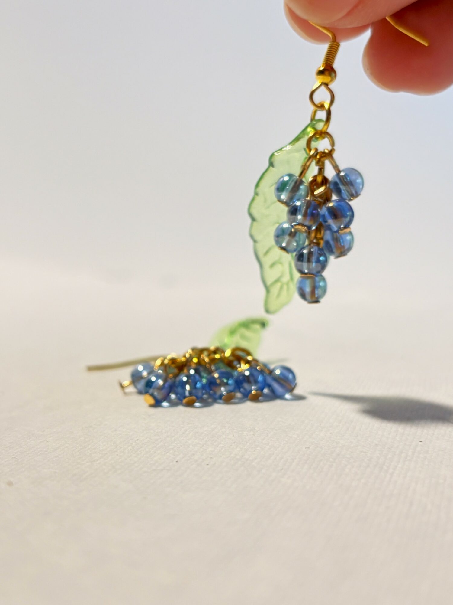 Hand holding a small earring with blue beads and a green leaf-shaped element, above a surface with similar beads. Word count: 20