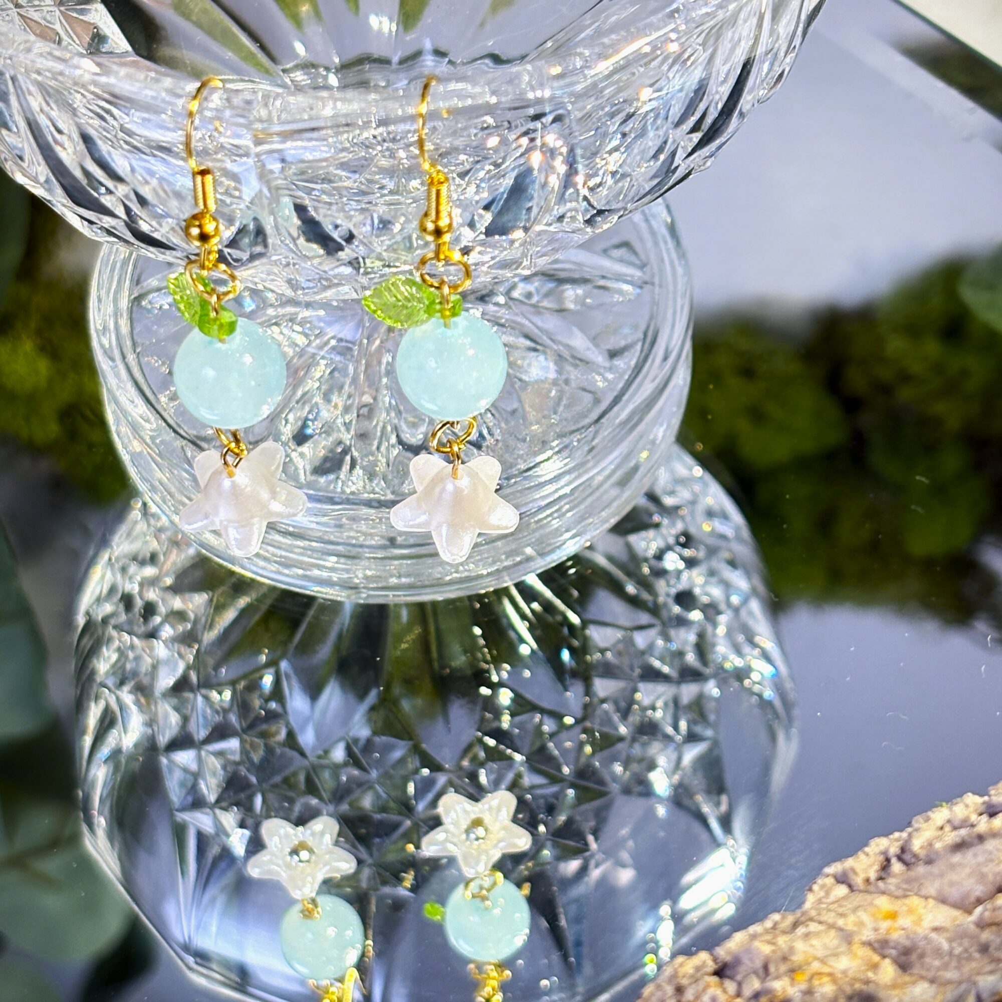 Pair of earrings with light blue beads, white flower charms, and gold hooks, hanging above a glass surface outdoors.