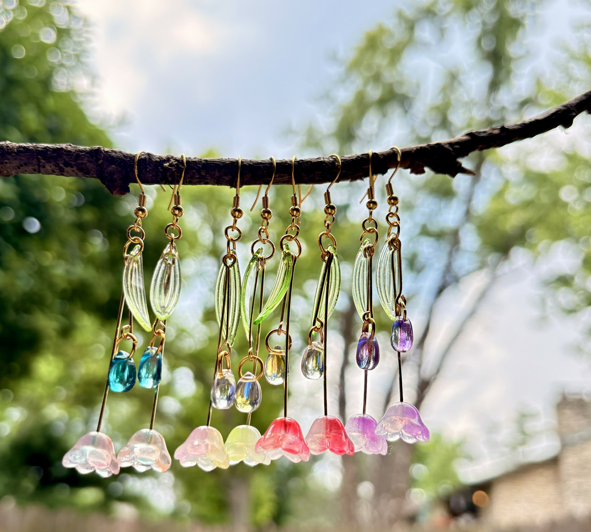 Seven pairs of colorful earrings hanging from a tree branch with a blurred outdoor background.