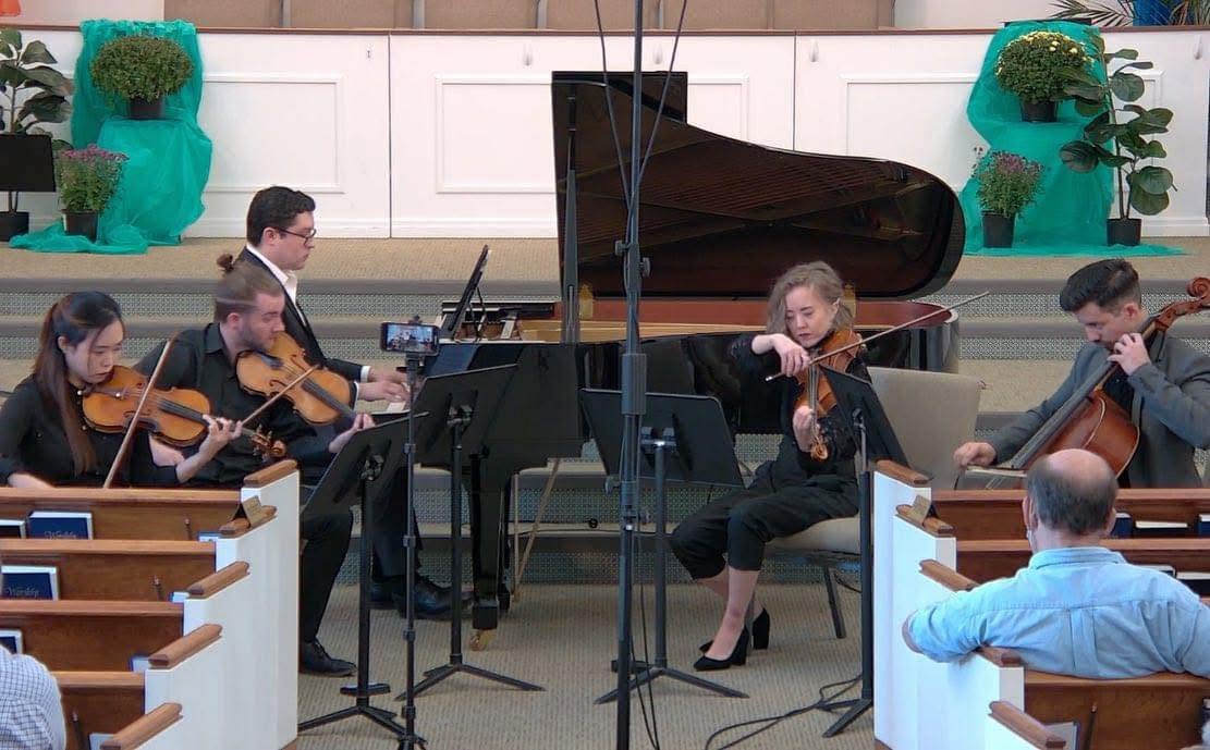 A group of musicians playing string instruments in a church with a grand piano in the background.