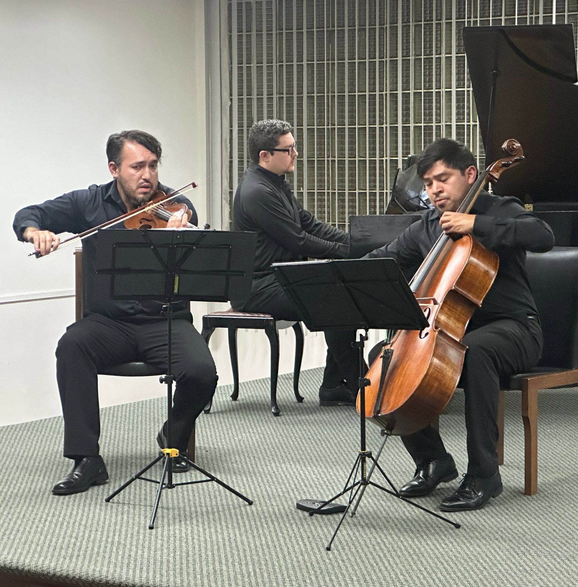 Three musicians perform with a violin, piano, and cello in a room with a barred window, music stands, and a grand piano.