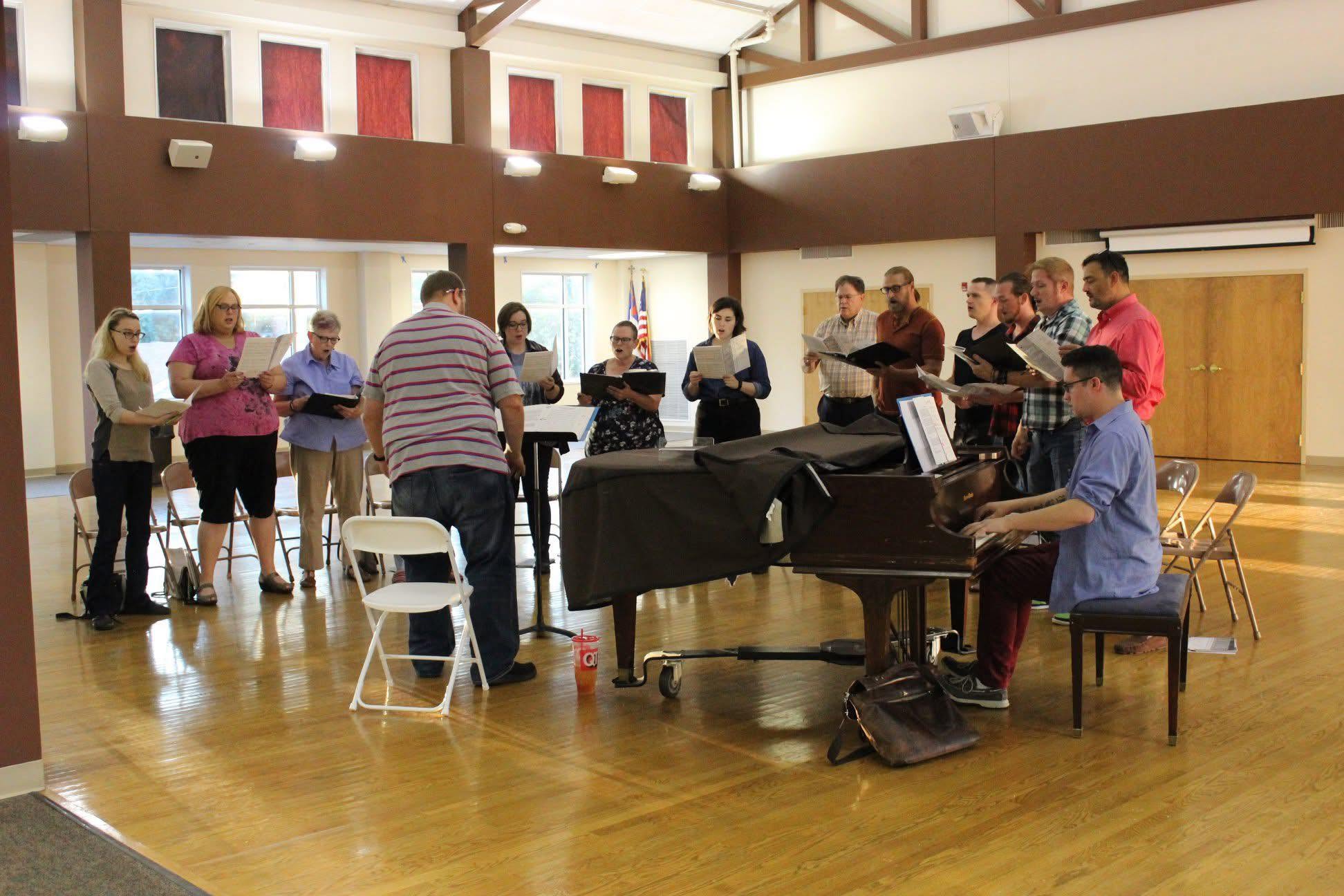 Group of people singing and playing piano in a spacious room with wooden floors and high ceiling.