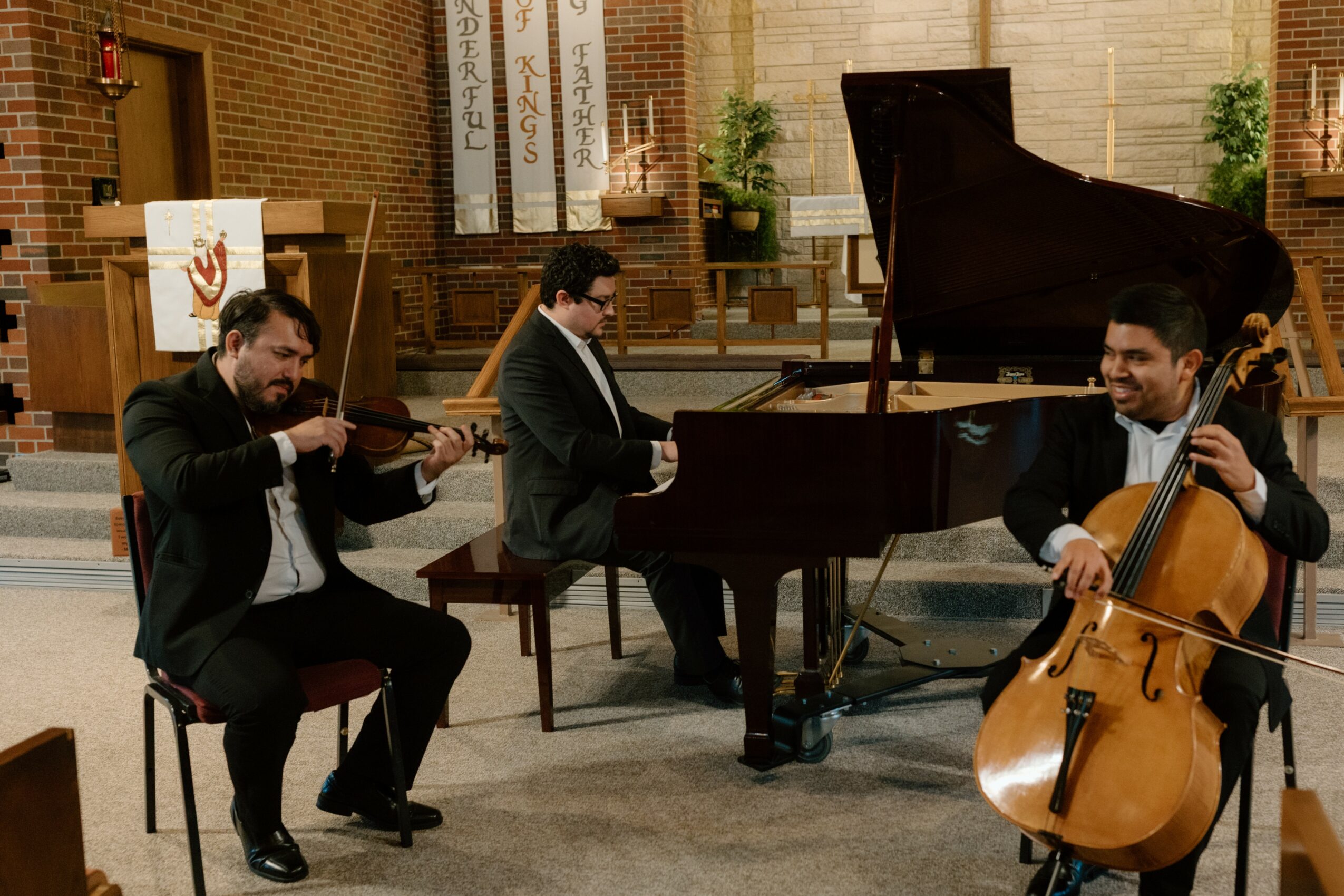 Three musicians perform in a church, playing violin, piano, and cello, with a brick wall and church interior in the background.