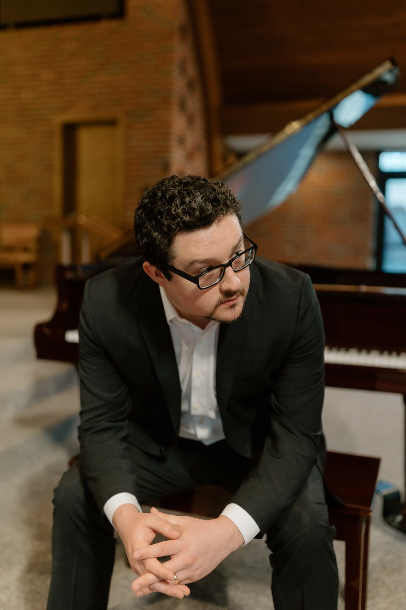 Man in black suit and glasses sitting on a piano bench in a music room, with a grand piano in the background.