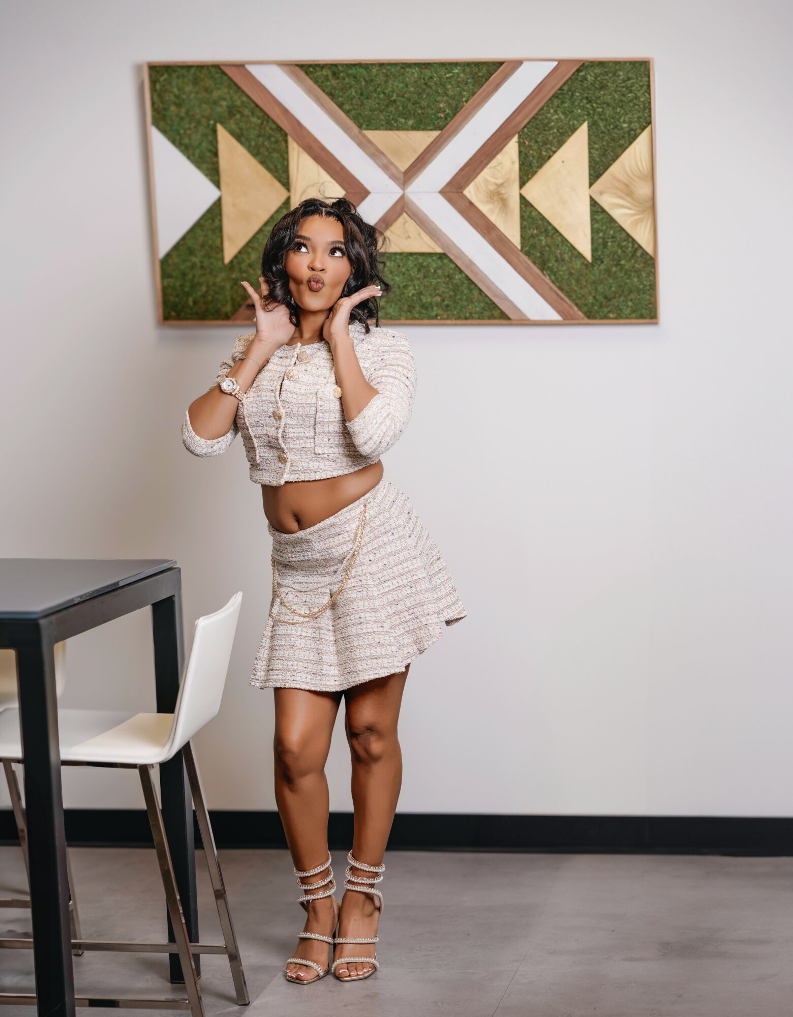 Young woman with curly hair in a white and beige outfit, standing in a room with a geometric wall art, a table, and a chair.