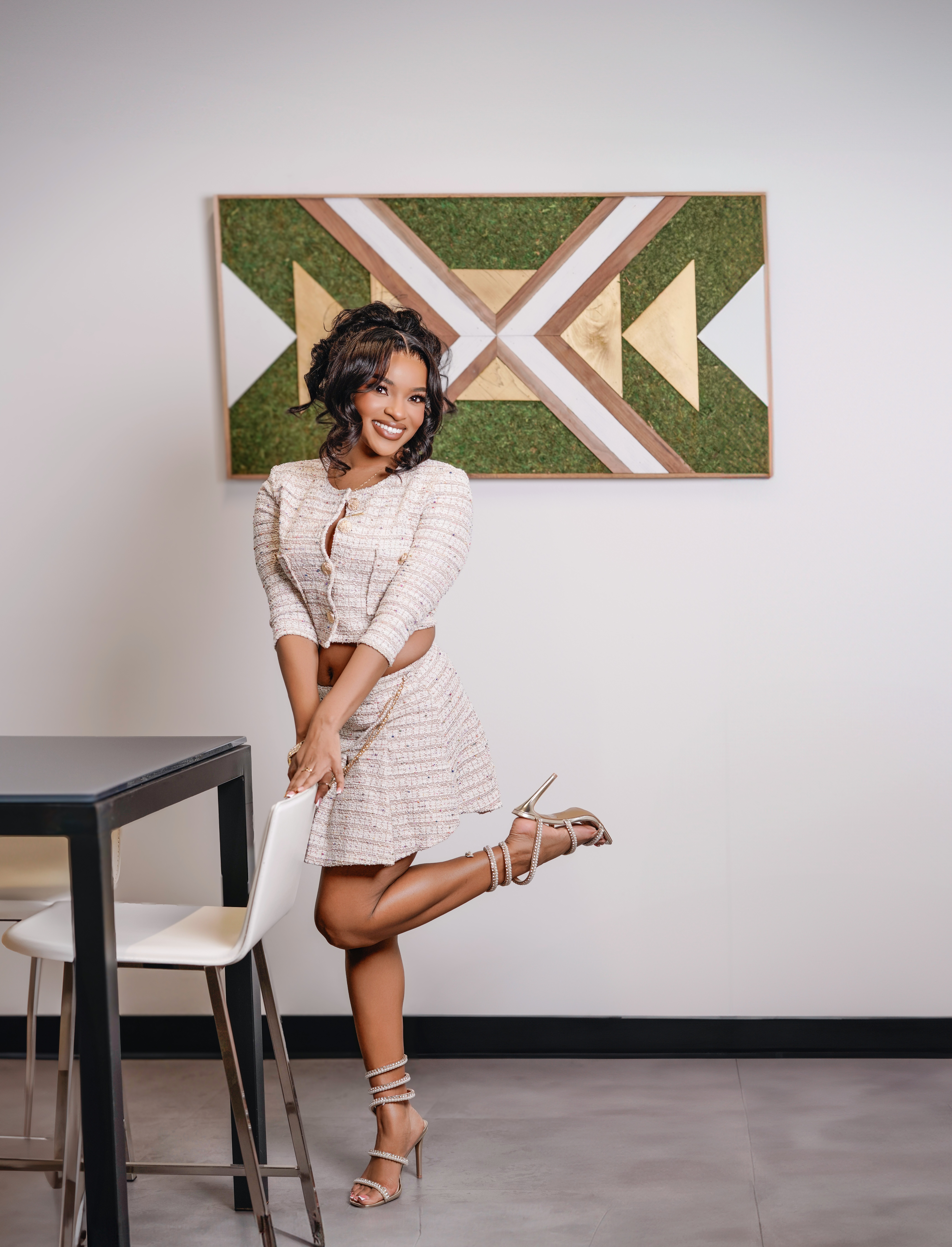 Young woman with curly hair in a white dress and heels, smiling, standing in front of a geometric wall art, one leg raised, holding her ankle.