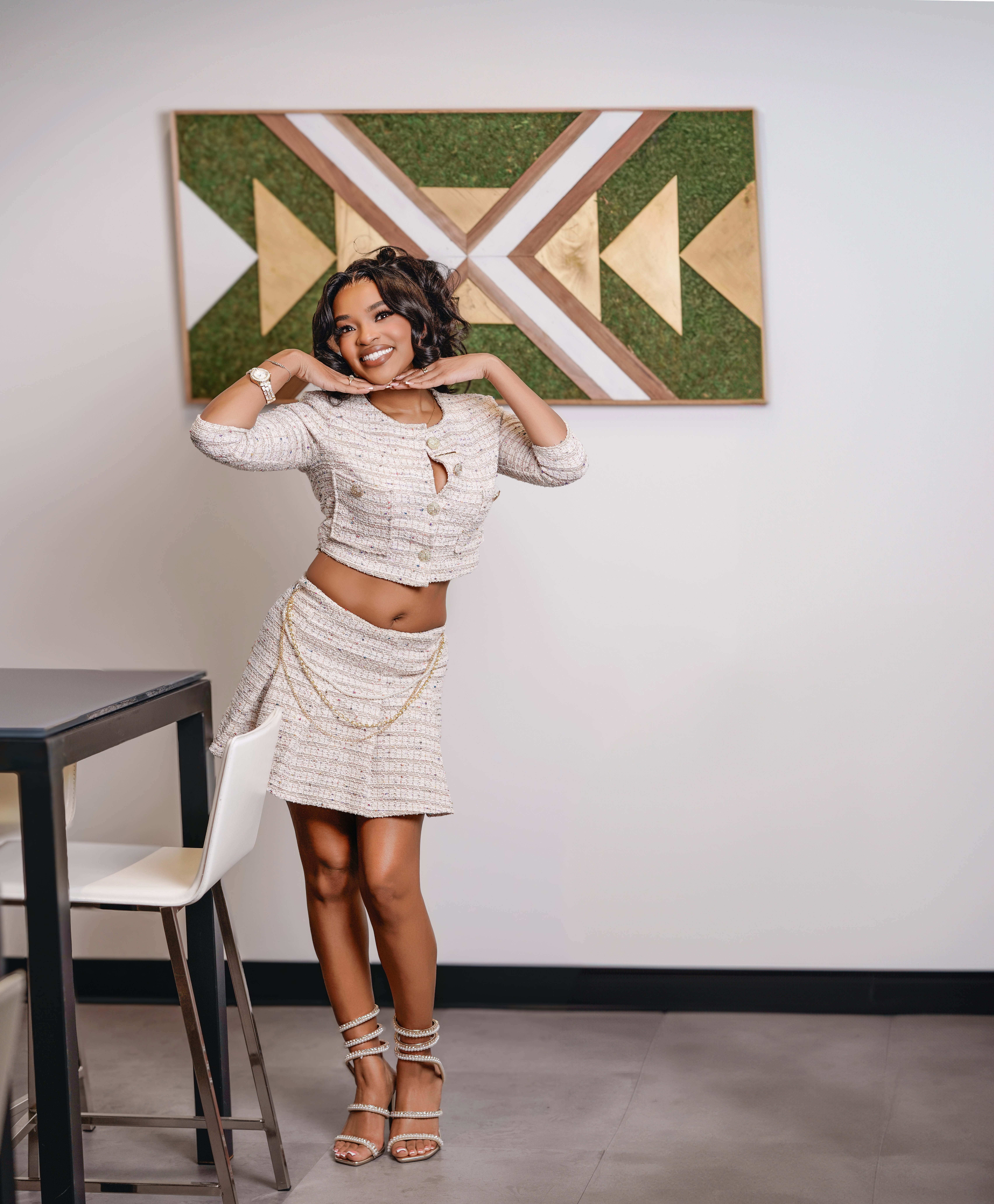 Young woman with curly hair smiling, standing in front of geometric wall art, wearing a striped skirt and crop top, heels, in a modern room.