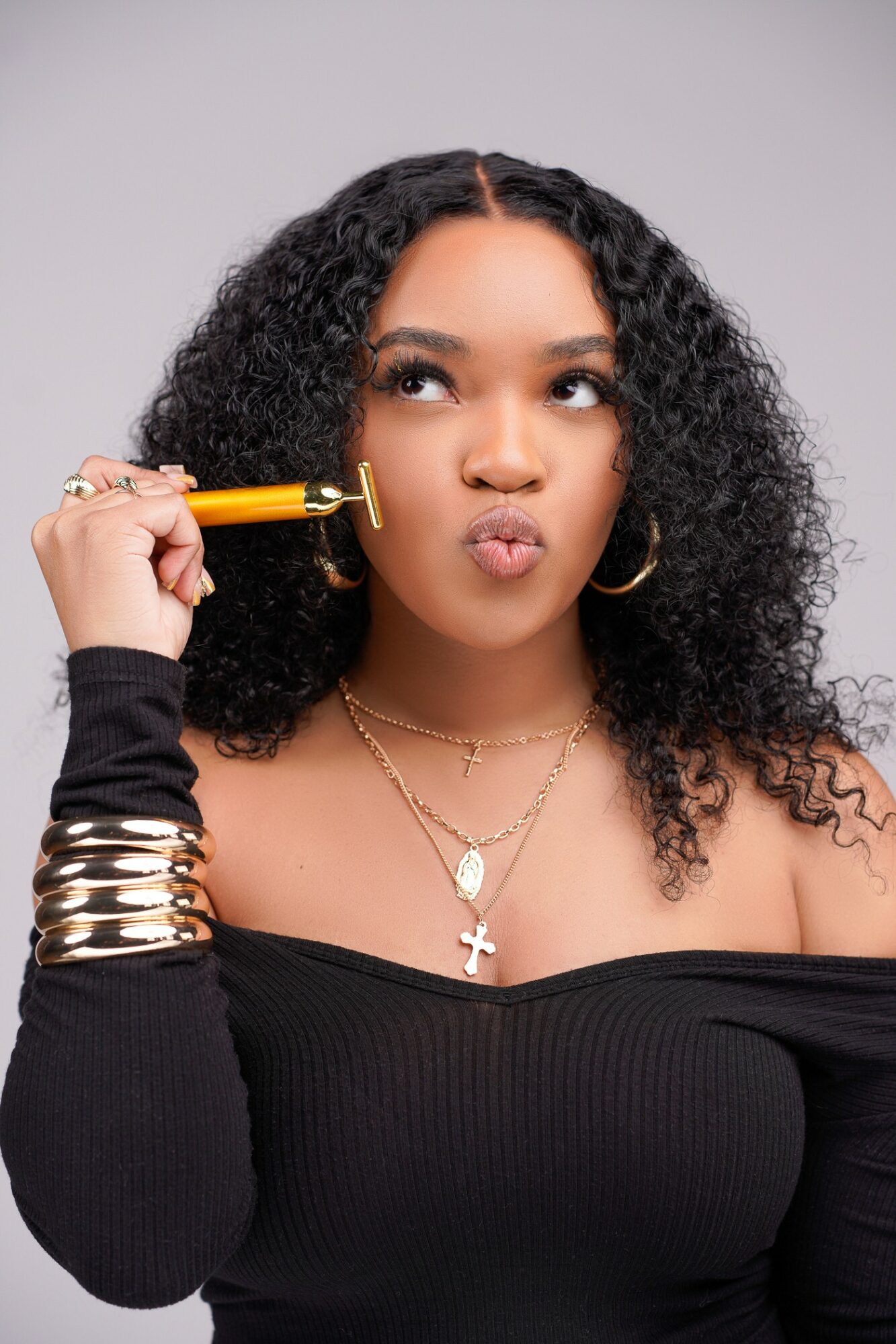 Young woman with curly hair holding a yellow object to her face, wearing jewelry and a black off-shoulder top.